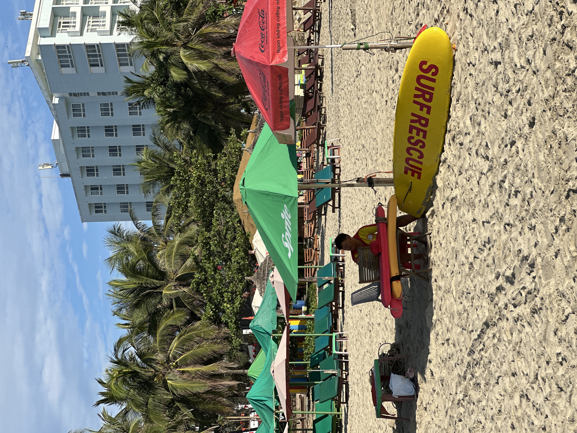 The image depicts a sunny day at a beach in Da Nang, Vietnam, likely My Khe Beach, known for its rows of palm trees and umbrellas. In the foreground, a lifeguard, wearing a red and yellow uniform, sits at a table under a red Coca-Cola branded umbrella. Beside him are two surfboards, one yellow with red "SURF RESCUE" lettering and another red. A collection of green and beige beach chairs and umbrellas are set up behind him. In the background, a multi-story light blue hotel building with numerous windows is visible, bordered by lush green palm trees. The sand is light beige and appears well-trodden with footprints.