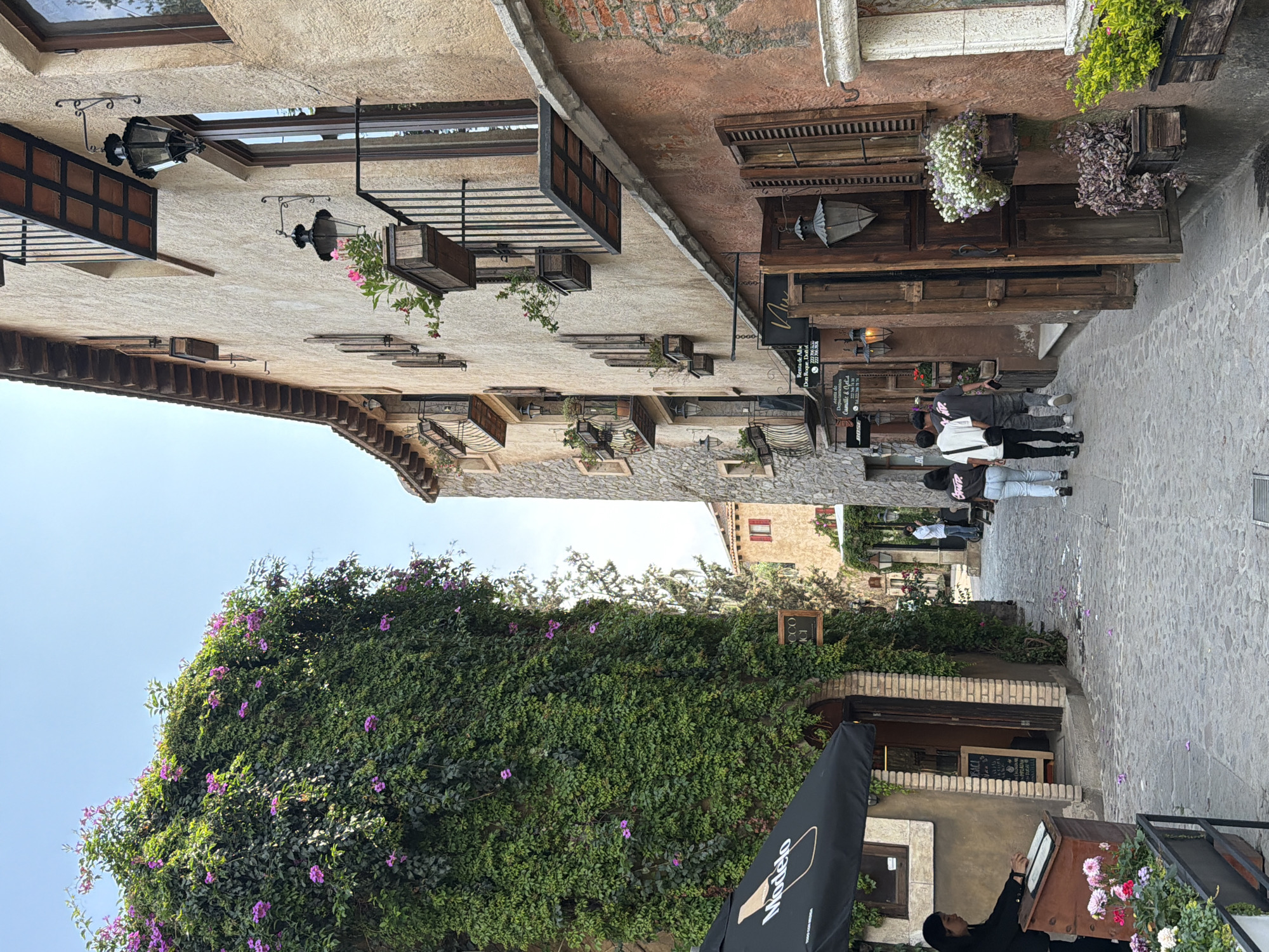 A street scene in Domingo Arenas, Mexico, shows a cobblestone street flanked by buildings with rustic architecture. The buildings display features such as wooden shutters, flower boxes, and decorative ironwork. One building is largely covered in a flowering vine. Several people are walking along the street. An outdoor cafe area is visible in the foreground, featuring a Modelo beer umbrella and flower arrangements.