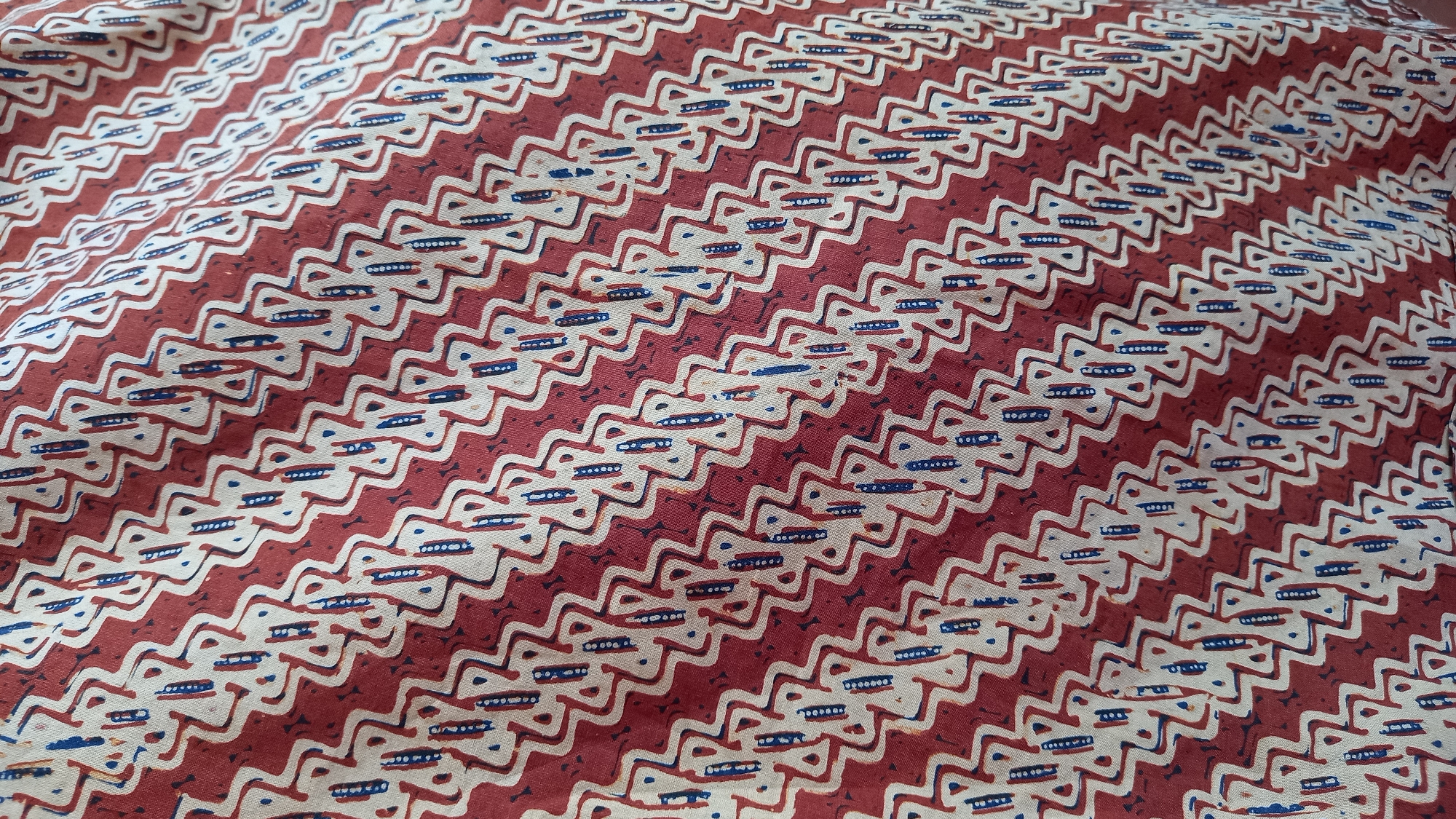The image displays a close-up, overhead view of a fabric with a repetitive zigzag pattern in white and red, interspersed with small blue dots and dashes. The fabric is likely a textile material, such as cotton or linen, and appears to be draped or folded, creating subtle shadows and highlights across its surface. The pattern is characterized by diagonal lines that zig and zag, forming a continuous, wave-like motif. The blue elements are small, oval shapes and short lines, accenting the white sections of the pattern. The overall impression is one of a decorative textile, possibly used for clothing, upholstery, or home decor. The image does not depict any specific location in South Tangerang, Indonesia, nor does it include any people, actions, or interactions.