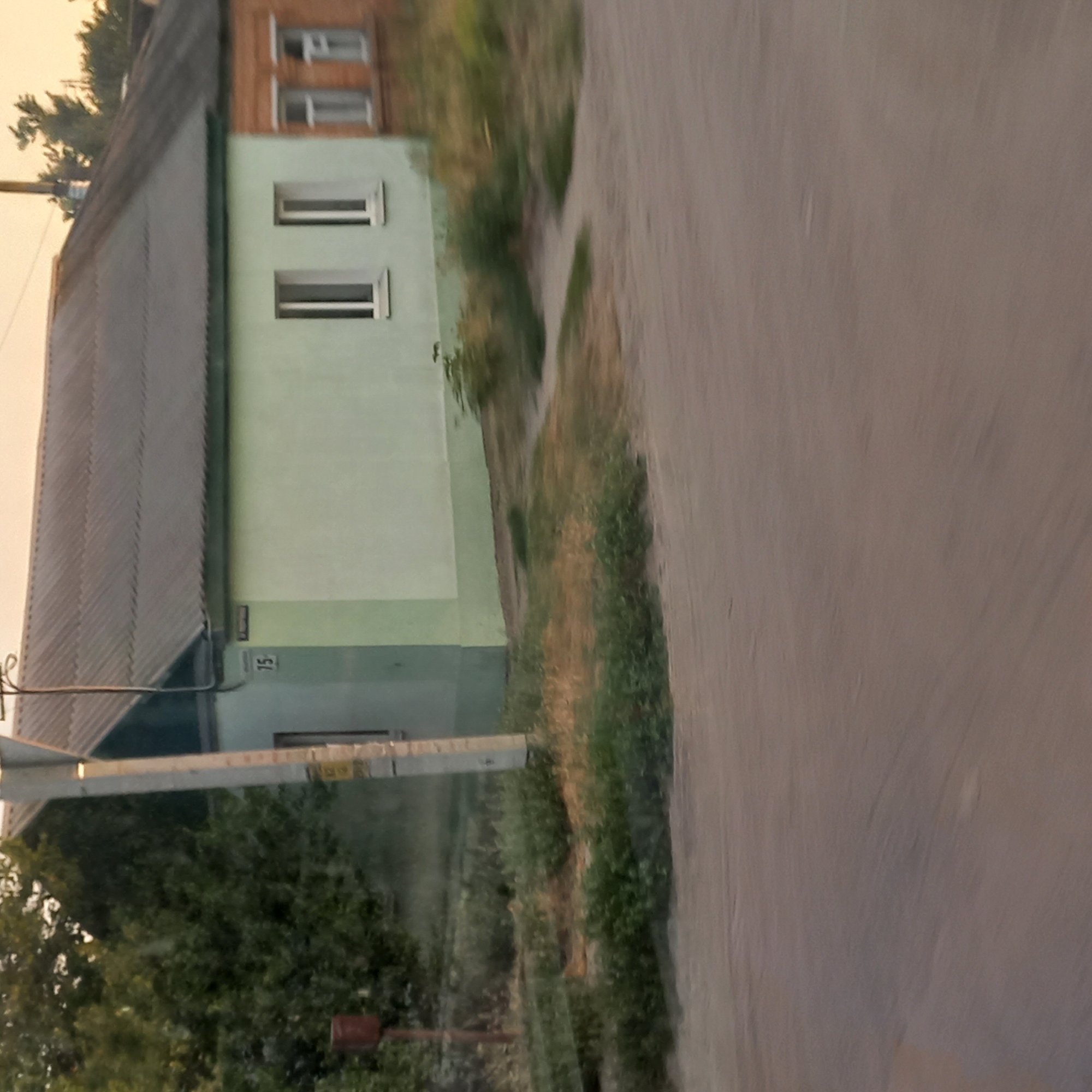 The image depicts a street view in Kropyvnytskyi, Ukraine, featuring a low-rise building with a light green facade and a section of orange brick. The building has a dark, corrugated roof. Several windows are visible, some with white frames. A sign with the number "15" is affixed to the green section of the building. Adjacent to the building, there is overgrown vegetation and grass. A utility pole stands in front of the building, and a small, reddish-brown cylindrical object is mounted on a pole to the left. The foreground is dominated by a dirt or gravel road, suggesting a rural or semi-urban setting. The lighting indicates it is either early morning or late afternoon, with a soft, warm hue to the sky. No people are present in the frame.
