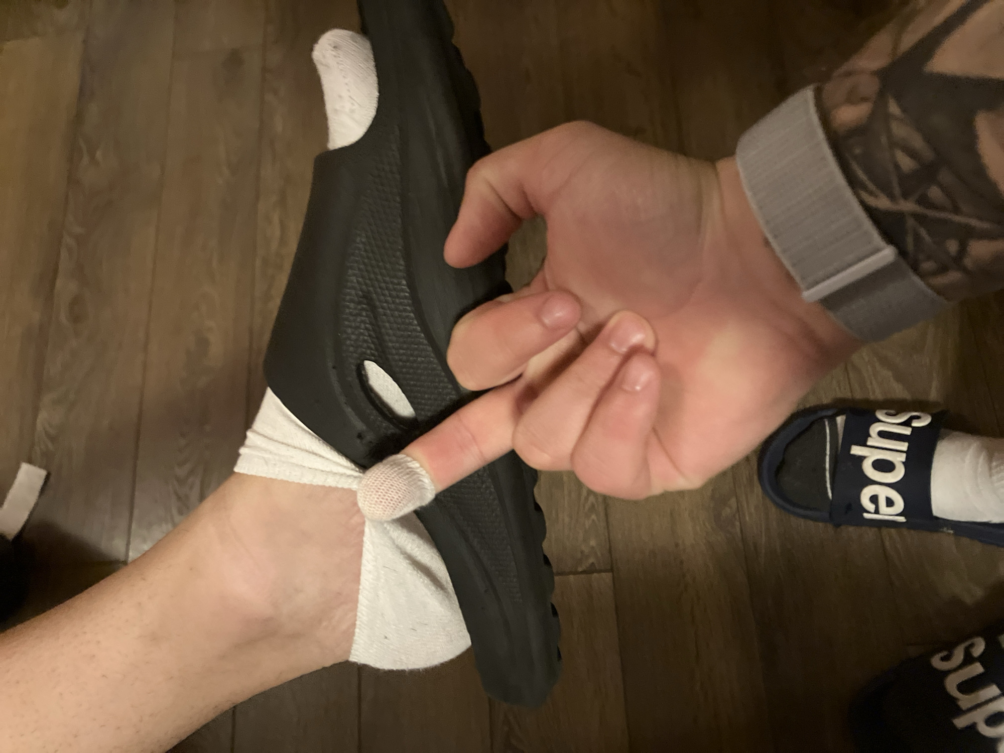 The image shows a close-up view of a person's foot wearing a white sock and a black slide sandal. A finger, with a band-aid on the tip, is touching the sandal. Another foot, also wearing a white sock and a navy blue slide sandal with the word "supe" visible, is in the lower part of the frame. The background appears to be a wooden floor. The image was taken indoors, likely in a residential setting. There is no information in the image to suggest it is located in Plovdiv, Bulgaria.