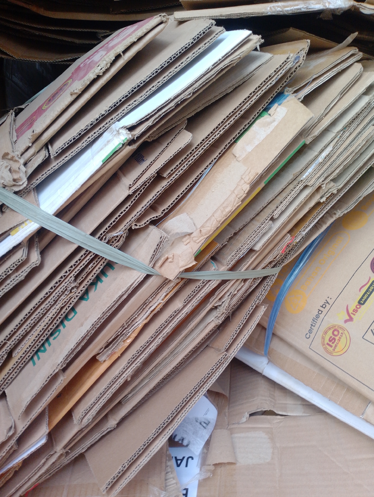 This is a close-up, overhead shot of a stack of used cardboard boxes. The boxes are layered in a slightly irregular fashion, with the corrugation of the cardboard clearly visible on the edges of many of them. Some of the boxes have printing on them, including what appears to be text in green and red, and an "ISO" certification logo. A green plastic strap is wrapped around a portion of the stack, holding it together. The lighting is somewhat diffuse, suggesting an indoor or shaded outdoor environment. The scene appears to be in a storage or recycling area, with the focus solely on the cardboard materials. There is no indication of the city of Batulicin, Indonesia in the image itself.