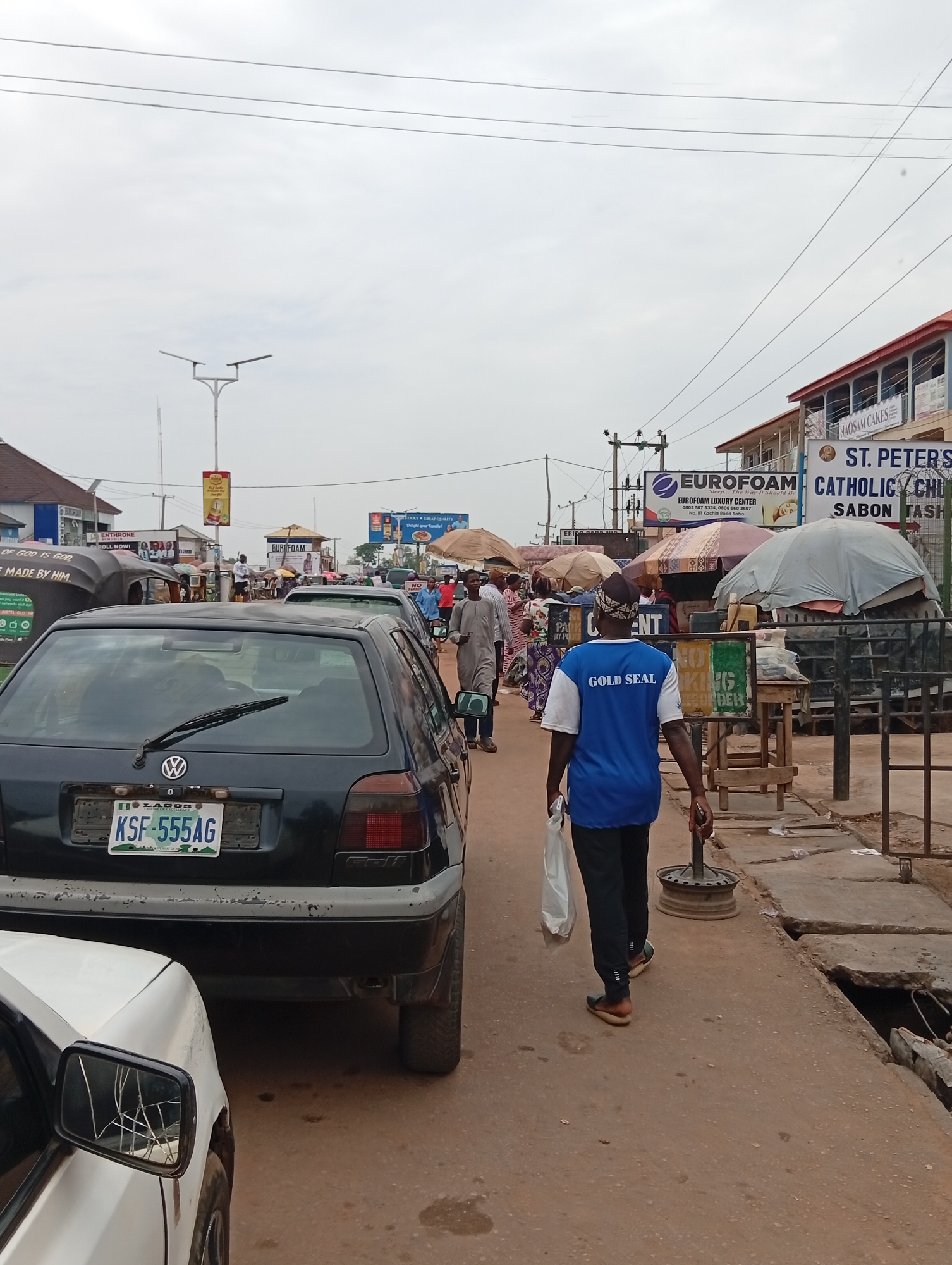 The image depicts a street scene in Sabon Tasha, Nigeria. A black Volkswagen Golf with license plate KSF-555AG is prominently featured. A man wearing a blue shirt with "GOLD SEAL" printed on it walks along the street, carrying a white plastic bag and holding a metal object. Other people are visible in the background, some under umbrellas. Several commercial signs are also visible, including "EUROFOAM" and "ST. PETER'S CATHOLIC CHURCH SABON TASHA". The road surface is cracked and broken, with visible damage. A white car is partially visible on the left side of the image, with a cracked side mirror. Overhead power lines are visible against a cloudy sky.