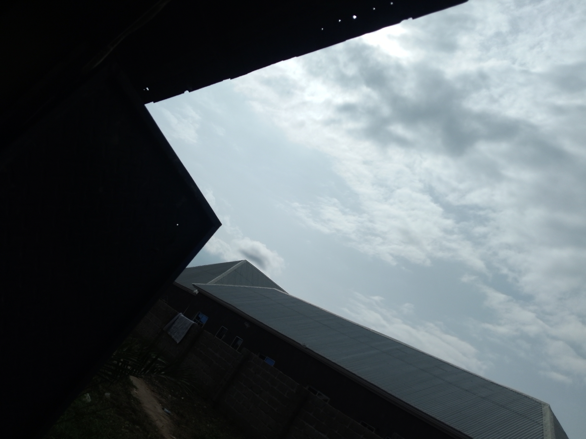 The image shows a low-angle view of a large, dark-colored building with a corrugated metal roof, viewed from what appears to be an interior space or a structure under construction. A portion of a black, angled wall or ceiling dominates the left side of the frame, obscuring the top left corner of the building. The building itself has a dark, brick-like base and a series of windows. A light-colored cloth is draped over a section of the brick wall. The sky above is overcast with clouds. The scene is set in an outdoor environment, with the ground appearing to be unpaved dirt or gravel in the foreground. The building's angled roof suggests a large industrial or commercial structure. The lighting is diffused due to the overcast sky, casting a muted light on the scene.