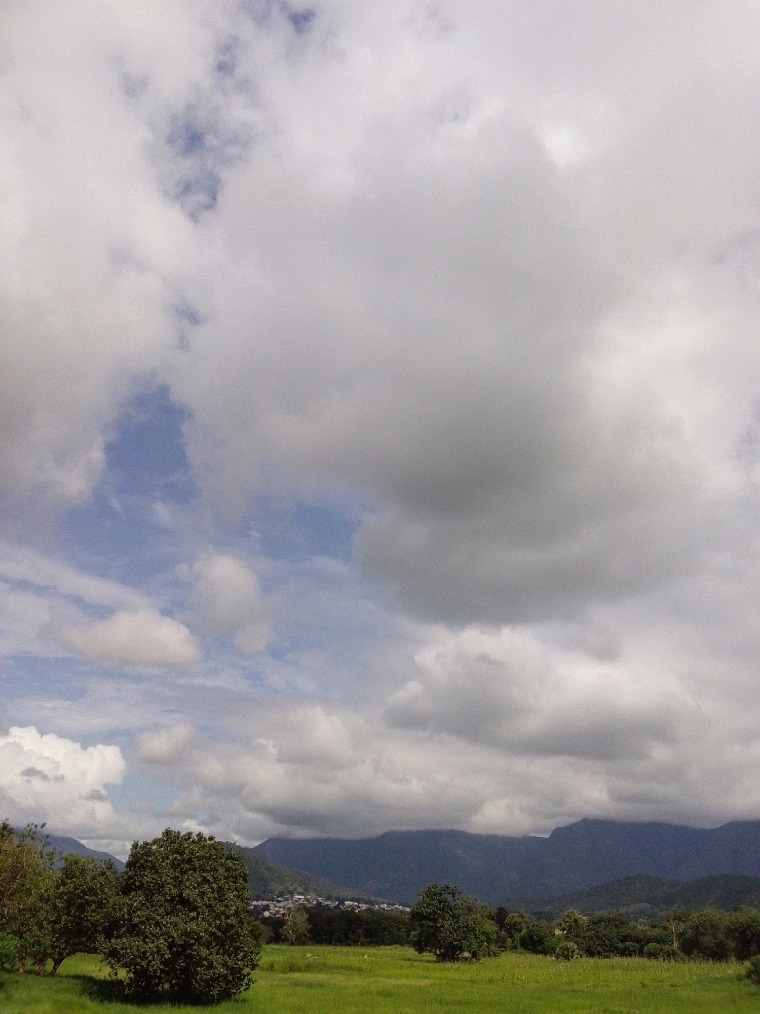 The image depicts a landscape scene with a focus on the sky and distant mountains. The sky is filled with a mix of white and gray clouds, with patches of blue visible. In the foreground, there is a grassy field with a few trees. In the middle ground, there is a town or village nestled amongst trees, and in the background, there are rolling hills and mountains. The overall impression is one of a natural, rural setting under a dynamic sky. The scene is likely located in or around Serti, Nigeria, given the context provided.