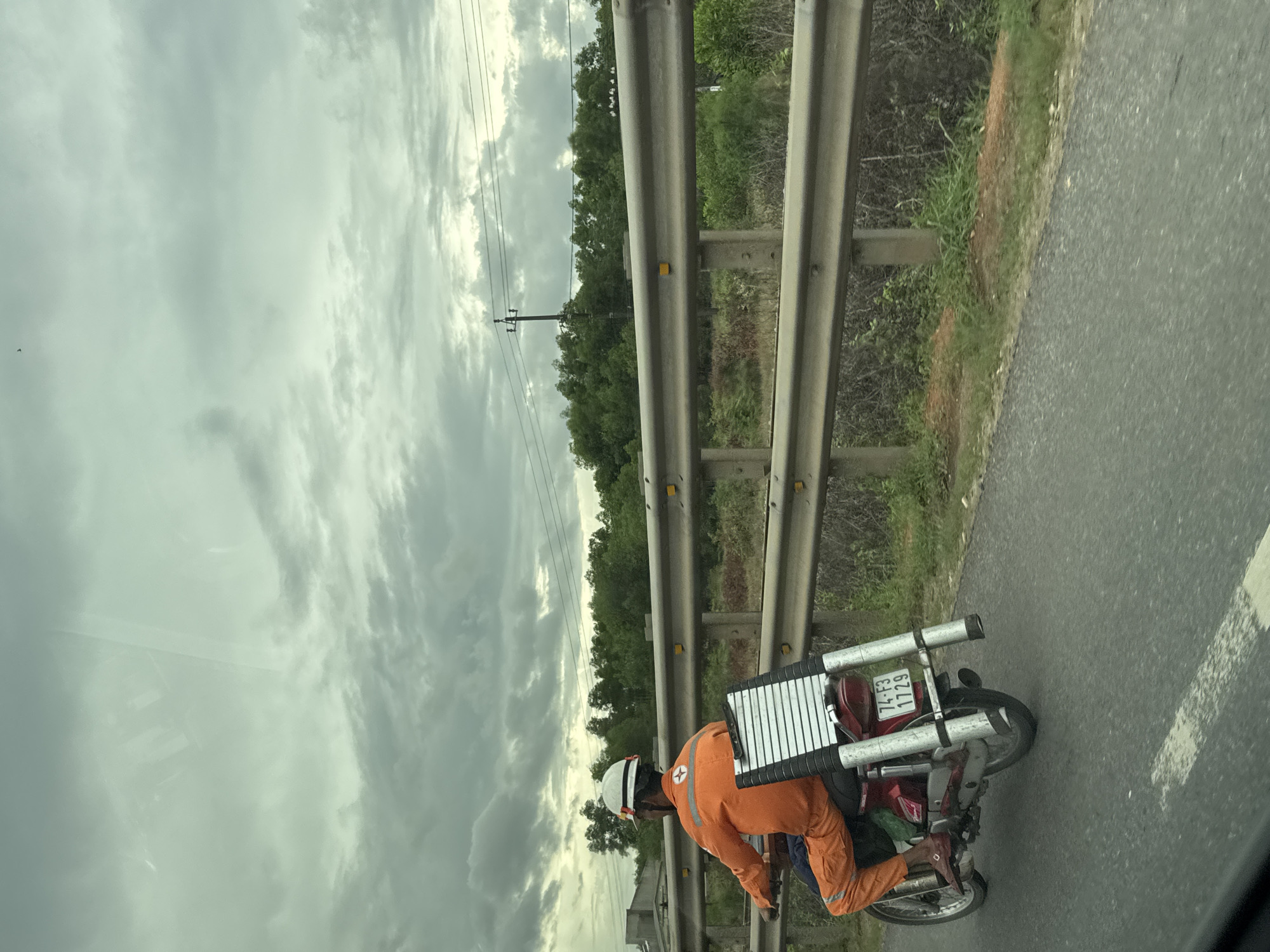 An individual wearing an orange uniform and a white helmet is riding a red motorcycle on a paved road in Hải Lăng Commune, Vietnam. The motorcycle is equipped with a silver ladder secured to the rear. The background features a guardrail, trees, utility poles, and a cloudy sky. The overall scene depicts a person commuting with equipment in a rural or semi-urban setting.
