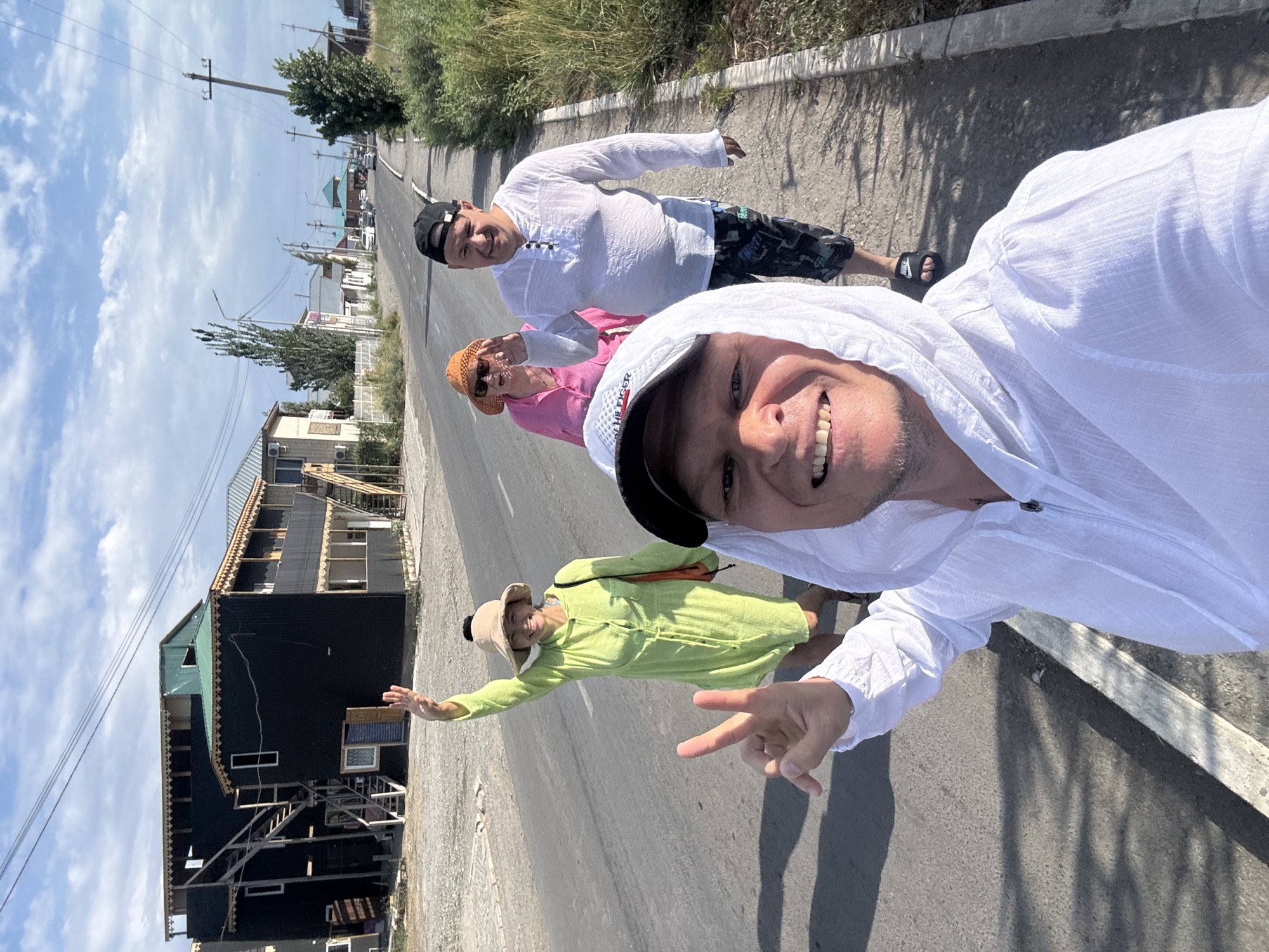 In Zharbulak, Kazakhstan, four individuals are captured in a selfie-style photograph on a paved road. The man in the foreground, wearing a white hooded jacket and a baseball cap, is smiling broadly and holding up two fingers in a peace sign. Behind him, a woman in a bright green dress and a wide-brimmed hat waves. Next to her, a woman in a pink shirt and an orange hat appears to be waving as well. The fourth person, a man in a white shirt and patterned shorts, is also visible, walking alongside the others on the sidewalk. The background features buildings with dark facades, some with external staircases, suggesting a residential or commercial area. Power lines and vegetation line the street, and the sky is partly cloudy. The scene conveys a casual, outdoor activity, possibly a stroll through the town.