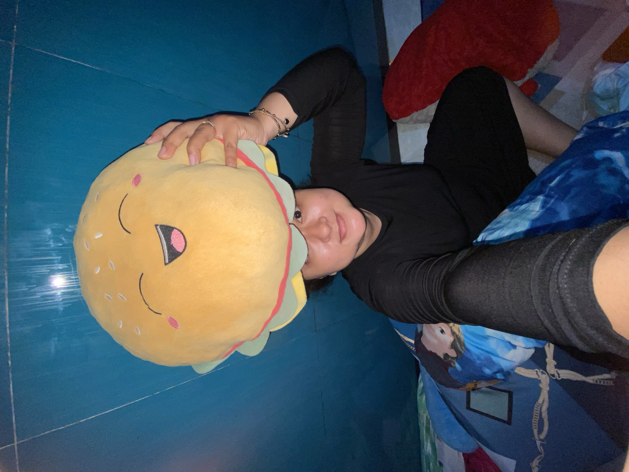 A person wearing black clothing is lying down on a bed covered with blue and white patterned bedding. A large, yellow, smiling burger plush toy is held above the person's head. The person is facing the camera, with a slight smile. The background appears to be a blue tiled floor. The location is presumably indoors in Gelam, Indonesia.