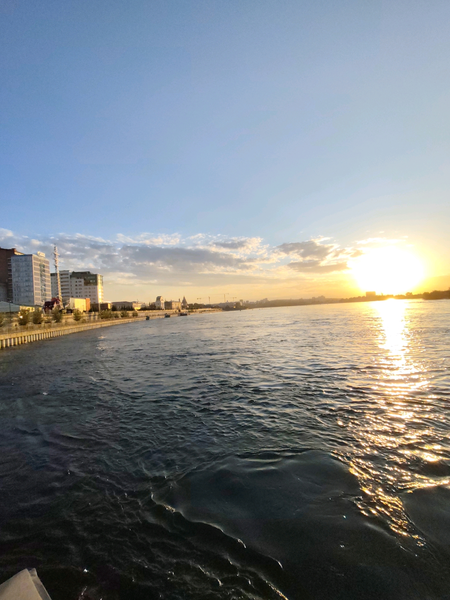 The image depicts a sunset over the Angara River in Irkutsk, Russia. The sun is low on the horizon, casting a warm golden glow across the water and illuminating the sky with hues of orange and yellow, mixed with shades of blue and white clouds. The river occupies the foreground and middle ground, with gentle waves reflecting the sunlight. Along the left bank, a city skyline is visible, featuring modern buildings, some with reflective glass facades. A promenade with trees and a decorative railing runs along the embankment. In the distance, more buildings and structures can be seen, including what appears to be a church steeple and construction cranes. The overall scene is serene and picturesque, capturing the natural beauty of the sunset over the urban landscape. There are no people clearly visible in the image.