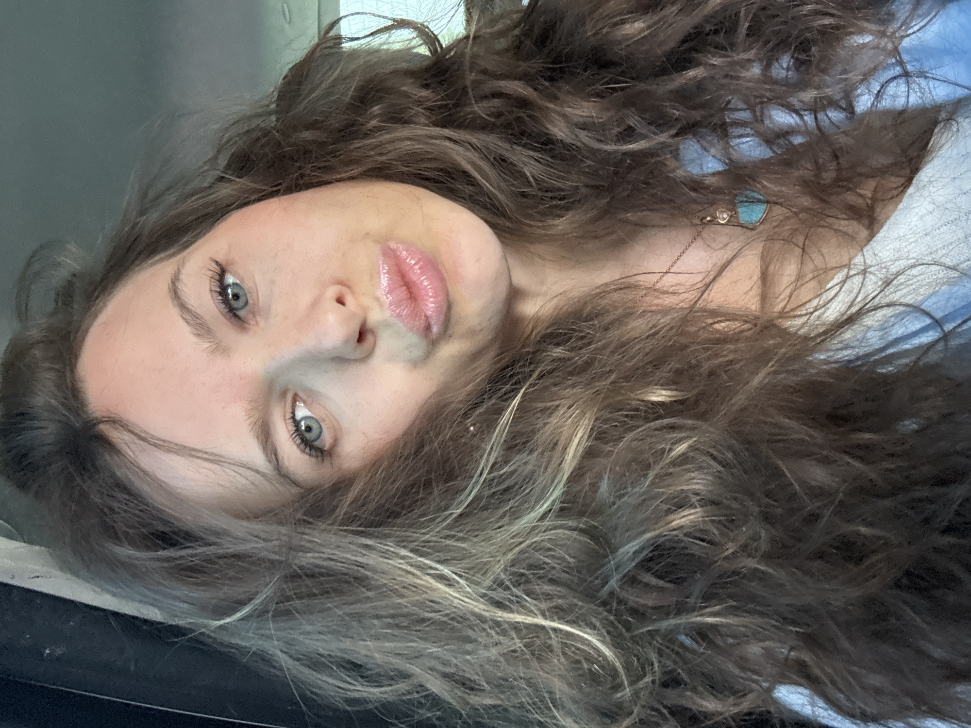 A close-up, rotated portrait shot captures a young woman looking directly at the viewer. Her head is tilted, and her long, wavy brown hair, featuring lighter blonde highlights, frames her face and spreads across the frame. She has striking blue eyes with defined brows and long eyelashes. Her lips are full, glossy, and a natural pink, slightly parted in a pout.
She appears to be wearing a light-colored, possibly blue or white, top, visible on the right side of the image. A delicate necklace adorns her neck, featuring a thin chain and a small, light blue, heart-shaped pendant, accompanied by another tiny clear stone or charm. The background is out of focus, showing indistinct, blurry shapes that suggest an indoor setting or inside a vehicle, with darker tones on the left and a lighter, possibly textured, area on the right. The lighting suggests daytime. There is no visible text in the image. No specific location cues for Moscow, Russia are discernible within the image itself.