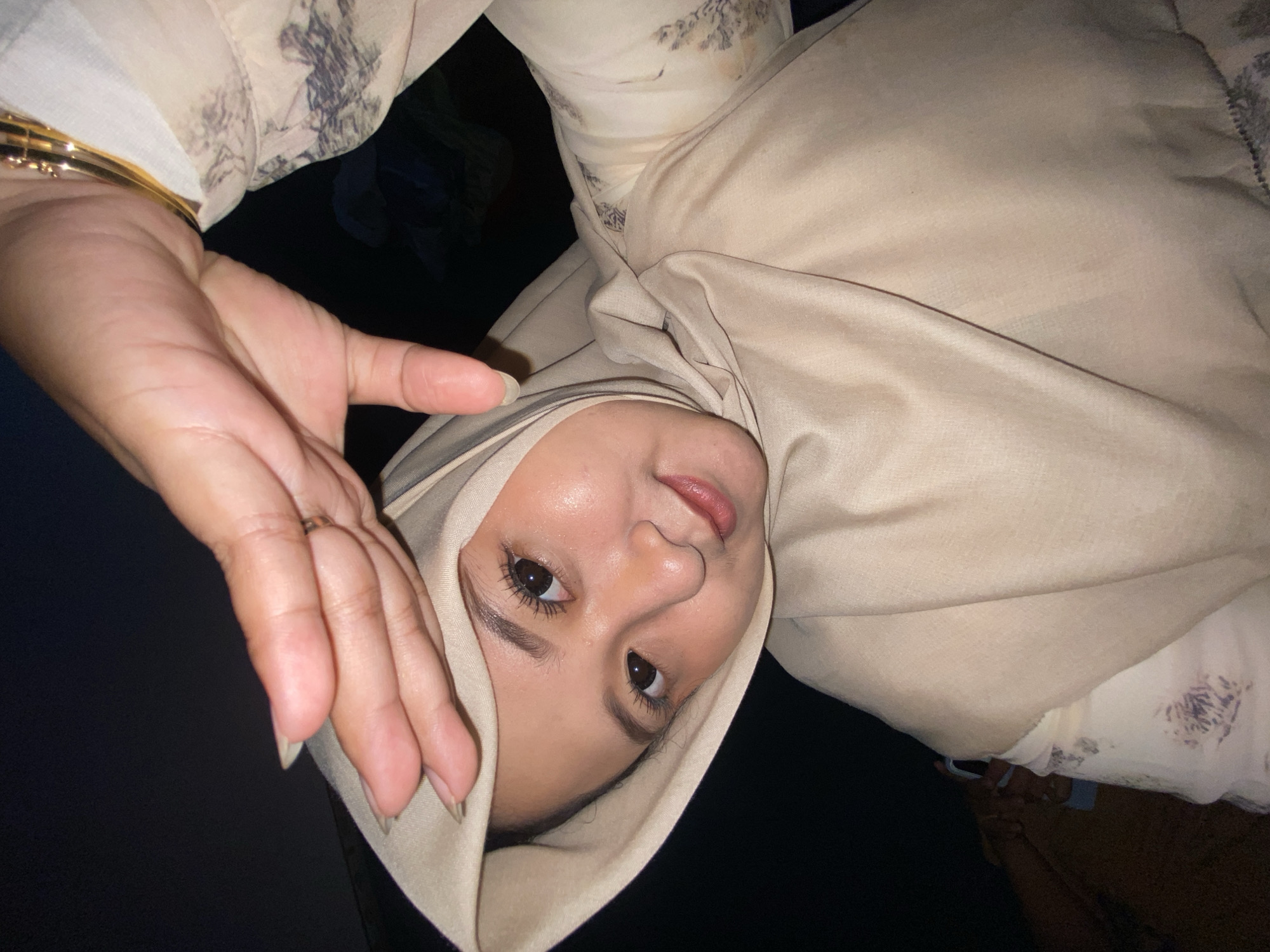 A woman wearing a beige hijab and a light-colored patterned blouse is seen in an upward-angled shot. Her right hand is positioned near her face, and a gold bracelet is visible on her wrist. The background is predominantly dark. The location, Gelam, Indonesia, is not evident from the image.
