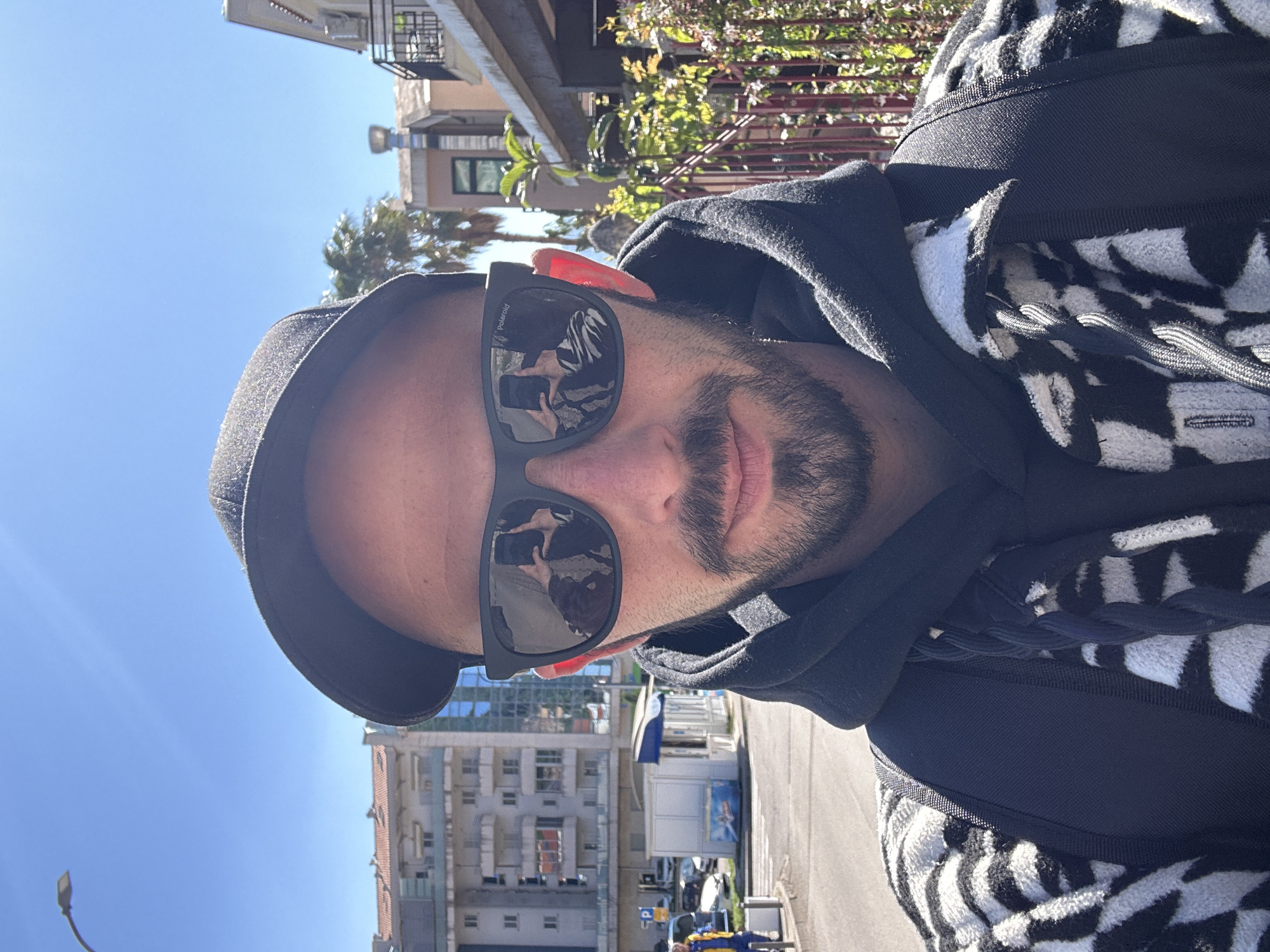 The image is a selfie of a man taken outdoors on a bright, sunny day in an urban or suburban environment, identified as Budva, Montenegro. The man is depicted from the chest up, looking directly at the camera. He is wearing a black baseball cap, black sunglasses with the brand "Polaroid" visible on the left lens, a black hooded sweatshirt, and an outer jacket or hoodie featuring a prominent black and white abstract or checkerboard pattern with a textured, fuzzy appearance. Drawstrings are visible on this outer garment.
The background shows a clear blue sky, indicating fair weather. Behind the man, multi-story buildings with balconies and windows line a street. Green foliage and trees are interspersed among the structures. A road with parked cars, a small white kiosk or booth, and a streetlamp are also visible. A partial parking sign, showing the letter "P", can be discerned in the lower background. The reflection in the man's sunglasses clearly shows the person holding a phone to take the picture. The scene conveys a casual, daytime moment.