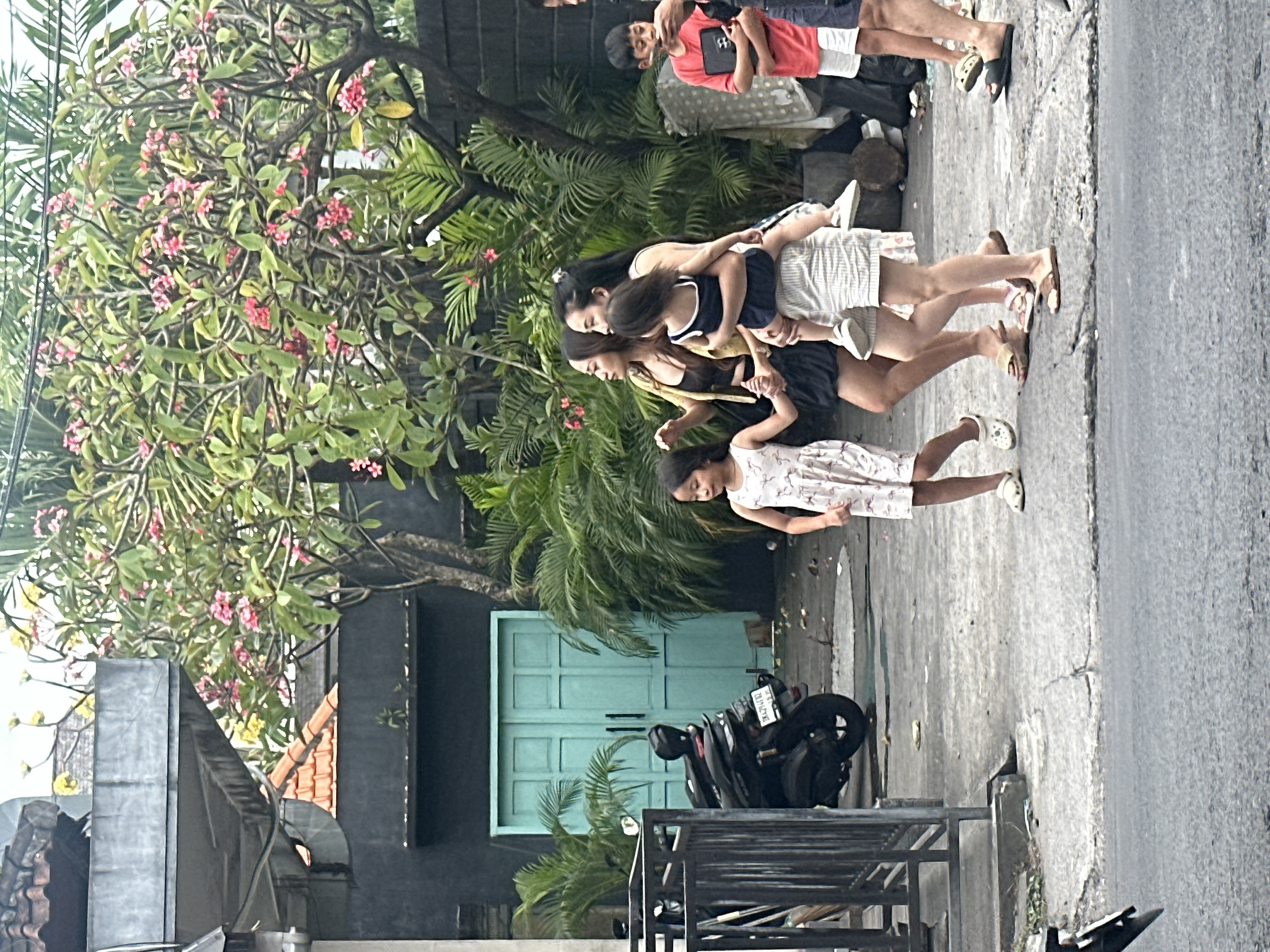 The image shows a group of people and a motorcycle on a street in North Kuta, Indonesia. In the foreground, a young girl in a white dress with a floral pattern stands on a cracked asphalt sidewalk, facing away from the camera. Behind her, a group of four individuals, appearing to be women and a younger girl, are posed closely together in a line. The woman in front is wearing a yellow top and dark skirt, with her arms around the person behind her, who is wearing a dark blue tank top and white shorts. The third person in line wears a light-colored top and white shorts, and the fourth person, who is largely obscured, appears to be wearing a dark top. The people are standing on a sidewalk in front of a building with a dark facade and a vibrant turquoise double door. Lush green foliage, including palm fronds and flowering trees with pink blossoms, surrounds the scene. A black motorcycle with a white license plate is parked on the sidewalk next to the turquoise door. In the background, the legs of two other individuals are visible, one wearing a red t-shirt and white shorts and the other wearing a dark shirt. The lighting suggests it is daytime, likely sunny. The overall mood appears casual and relaxed. No visible text other than the motorcycle's license plate, which is illegible.