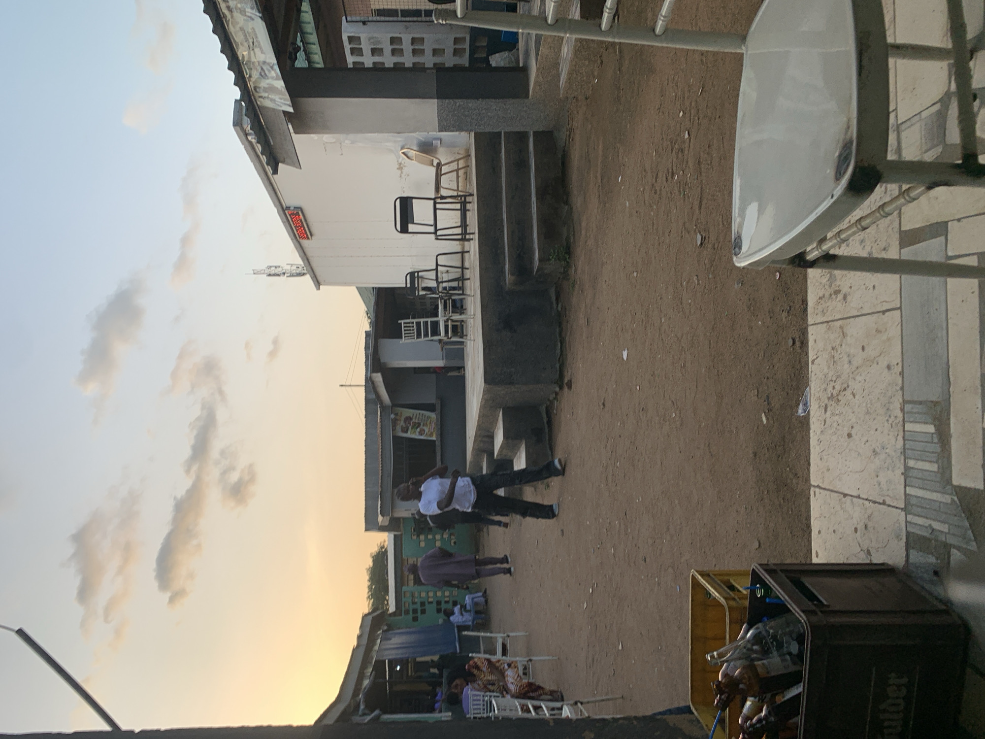 The image captures an outdoor communal area in the city of New Stadium, Nigeria, under the soft, transitioning light of either dusk or dawn. The ground is predominantly unpaved reddish-brown earth, with sections of tiled paving near the foreground and surrounding structures.
In the midground, a man wearing a white t-shirt and dark pants walks towards the viewer, appearing to be on a phone. Behind him, another man in a dark, possibly purple, traditional garment walks away. To the far left, a woman in a patterned dress sits on a white event chair.
On the right side of the scene, a prominent white single-story building features a red LED display sign on its facade and a cellular antenna mast on its roof. In front of this building, a raised concrete platform with three steps serves as a staging area, holding several chairs including two black metal chairs, a white event chair, and a gold-colored chair. To the left of the white building are smaller, simpler structures, one with a corrugated metal roof and a green and blue patterned wall, and another with a visible poster.
In the immediate foreground on the left, a brown plastic crate filled with various empty glass bottles, primarily beer or soda bottles, rests on a paved surface, partially obscuring a yellow plastic crate behind it. A white metal chair, partially cropped, is visible in the far right foreground. A thin, dark pole or mast is partially visible along the far left edge of the frame. The sky above displays a gradient of soft orange and blue hues with scattered, light clouds.