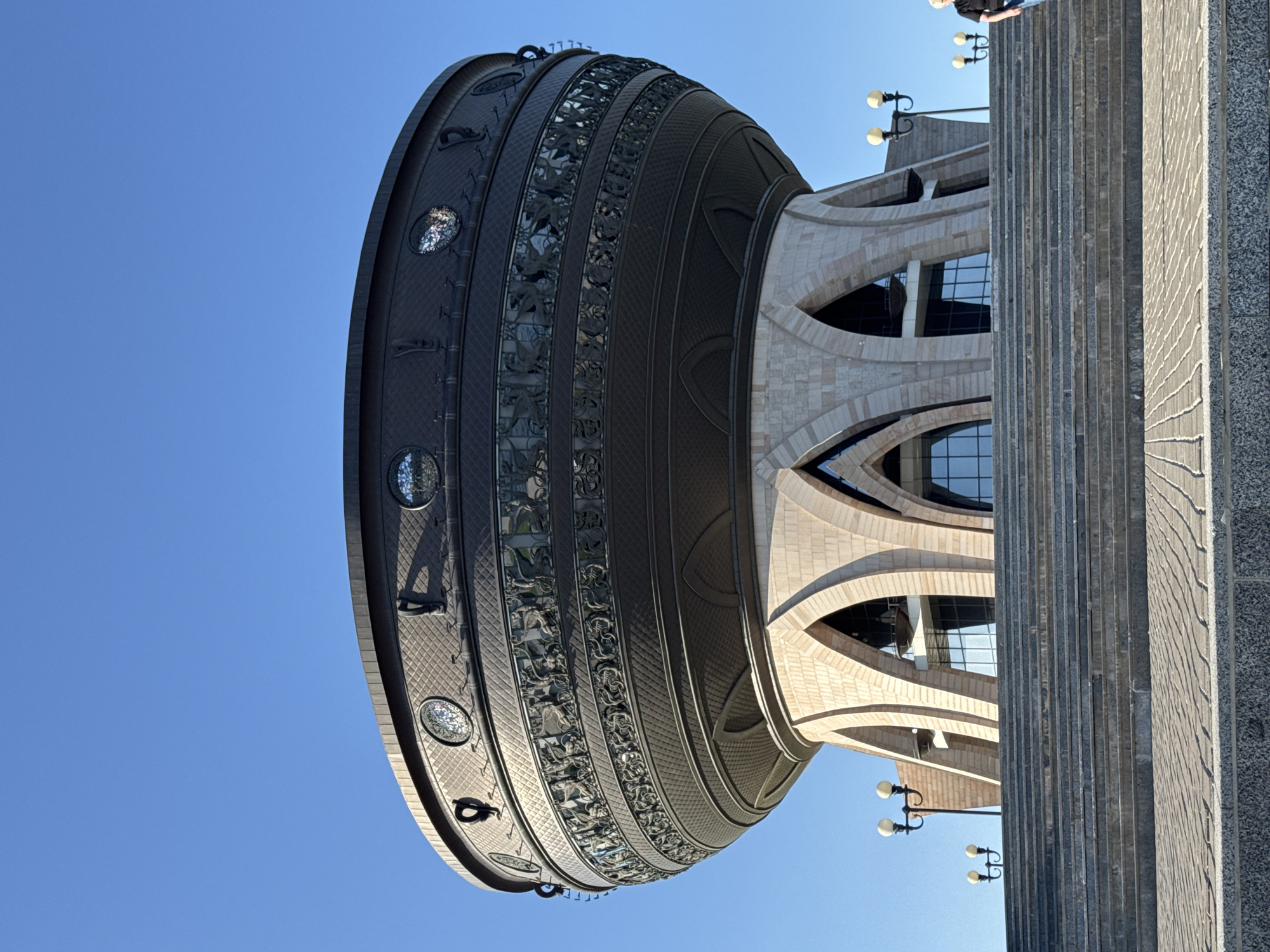 The image captures the Kazan Family Center, a distinctive building in Kazan, Russia. The structure features a large, stylized cauldron-shaped dome, decorated with circular windows and detailed relief patterns. The building's façade is light-colored, with a pattern of arched windows and curvilinear structural elements. A textured, gray pillar stands adjacent to the building, casting a shadow on the right side. Street lamps are visible near the base of the building, set against a clear, blue sky. Part of a person is visible at the upper right. The photograph is taken from a low angle, emphasizing the building's height.