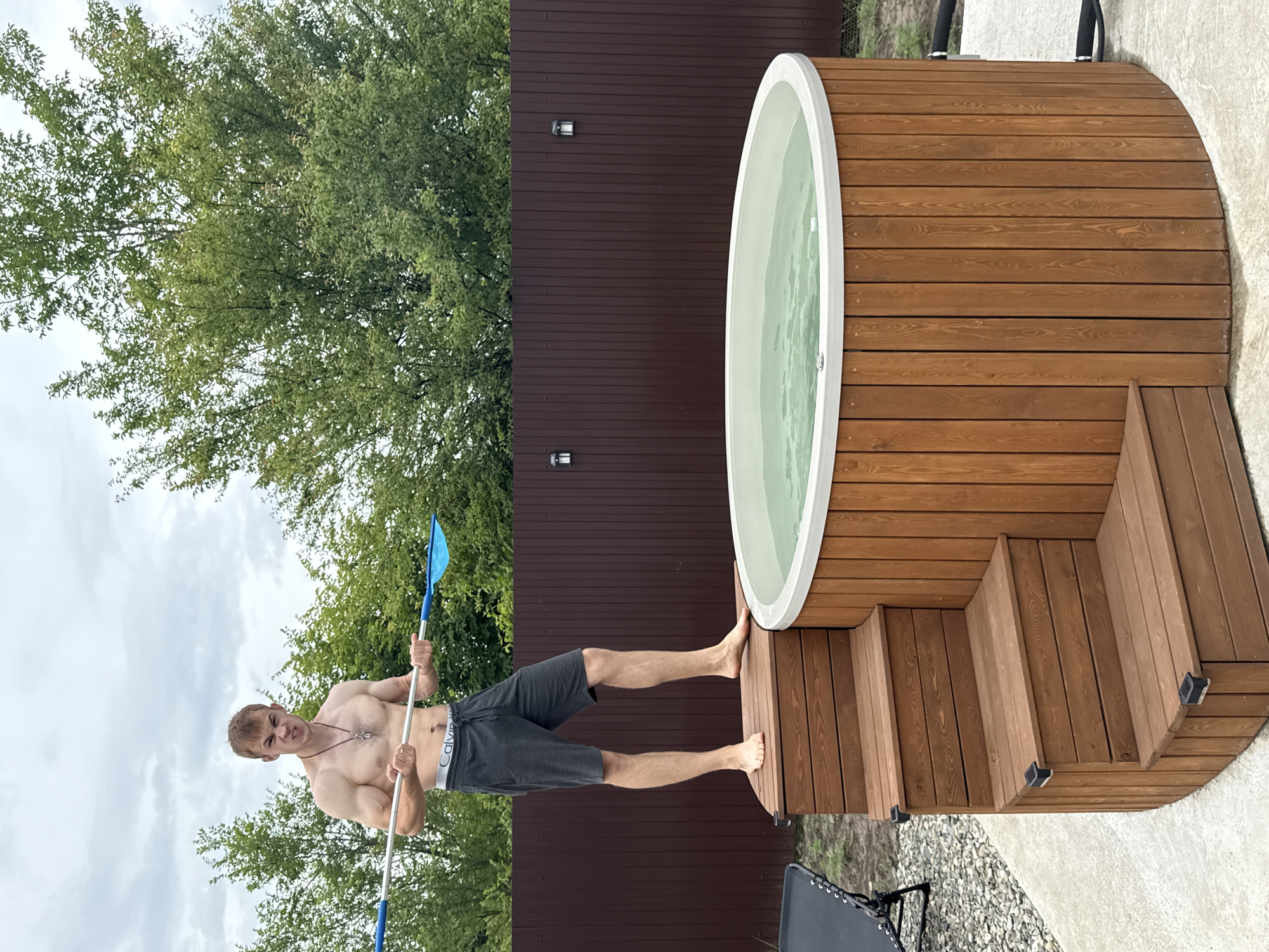In Starokorsunskaya, Russia, a shirtless man with dark hair stands on wooden steps next to a circular hot tub. He is wearing grey athletic shorts with the waistband of Calvin Klein underwear visible. The man holds a long pole with a blue net attachment, facing the camera with a grimace. The hot tub is filled with light green water and surrounded by wooden planks. Behind the hot tub is a dark brown wooden fence with two outdoor lights mounted on it. Lush green trees and an overcast sky form the background. To the left of the steps, a dark chair is visible on a gravel surface.