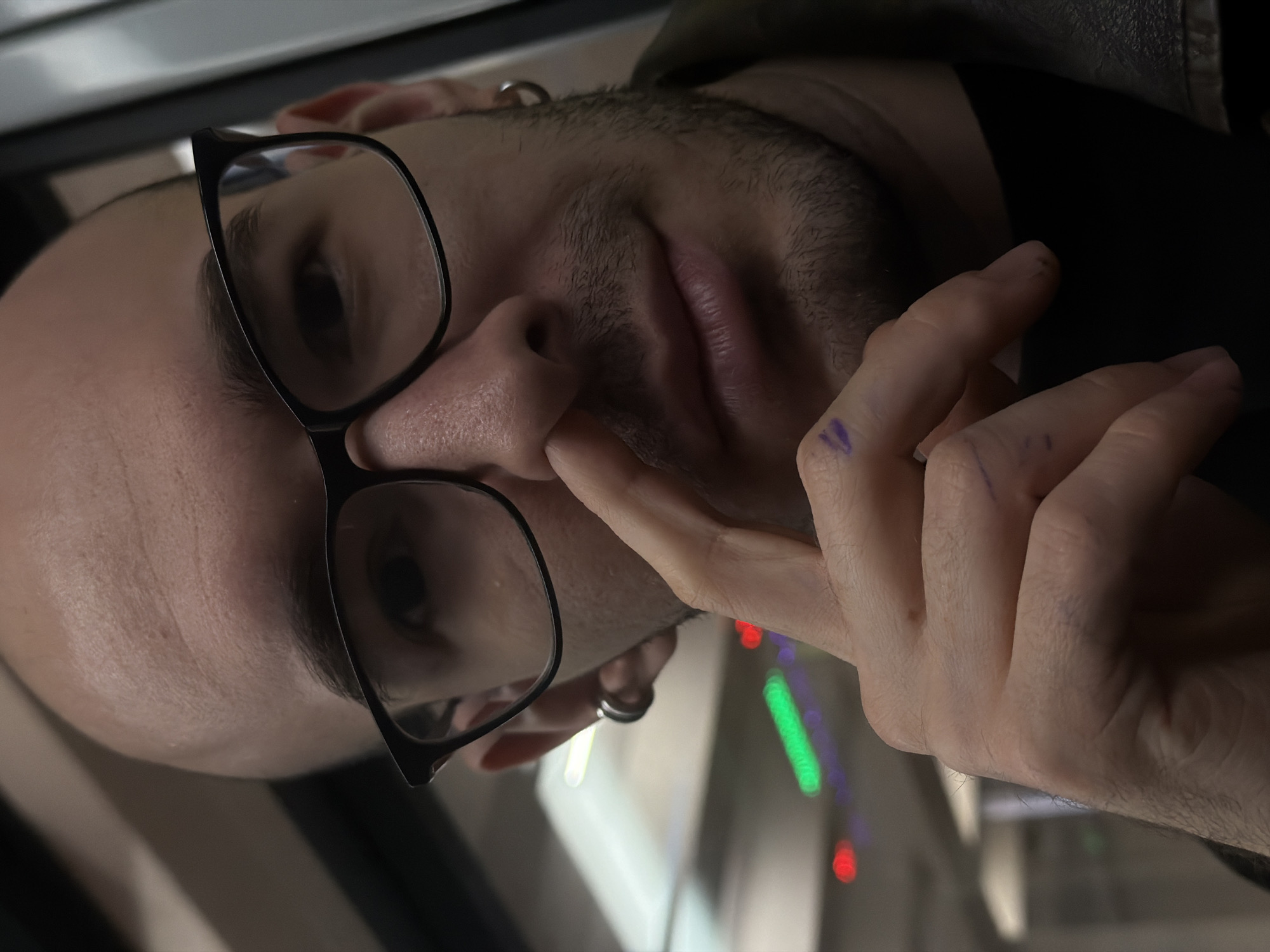 A close-up, low-angle shot depicts a person wearing glasses and a dark jacket. The person's hand is positioned near their nose, with fingers slightly bent. The background is blurred and contains vertical streaks of green, red, and purple lights, suggesting an urban setting, although the location is uncertain. There is a visible earring on the person's ear. There is some markings on the hand.