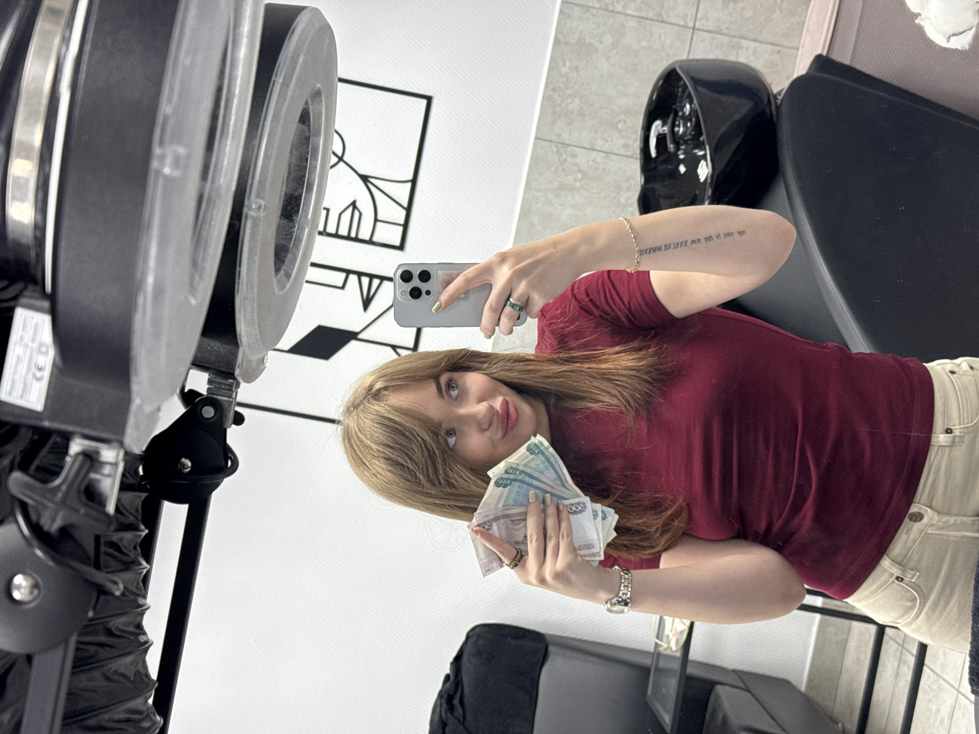 A young woman with shoulder-length brown hair and bangs is taking a selfie in what appears to be a salon or barbershop. She is wearing a maroon t-shirt and beige pants. She is holding a fan of Russian rubles in her left hand and is taking the photo with her right hand, which is holding a silver smartphone. She is wearing a watch on her left wrist and several rings on her fingers. A tattoo is visible on her right forearm. In the background, there is a black hair salon chair, a sink, and a large piece of salon equipment suspended from the ceiling. The walls are white and decorated with abstract line art. The floor is tiled. The image is taken from a slightly low angle, with the woman's face prominent in the foreground.