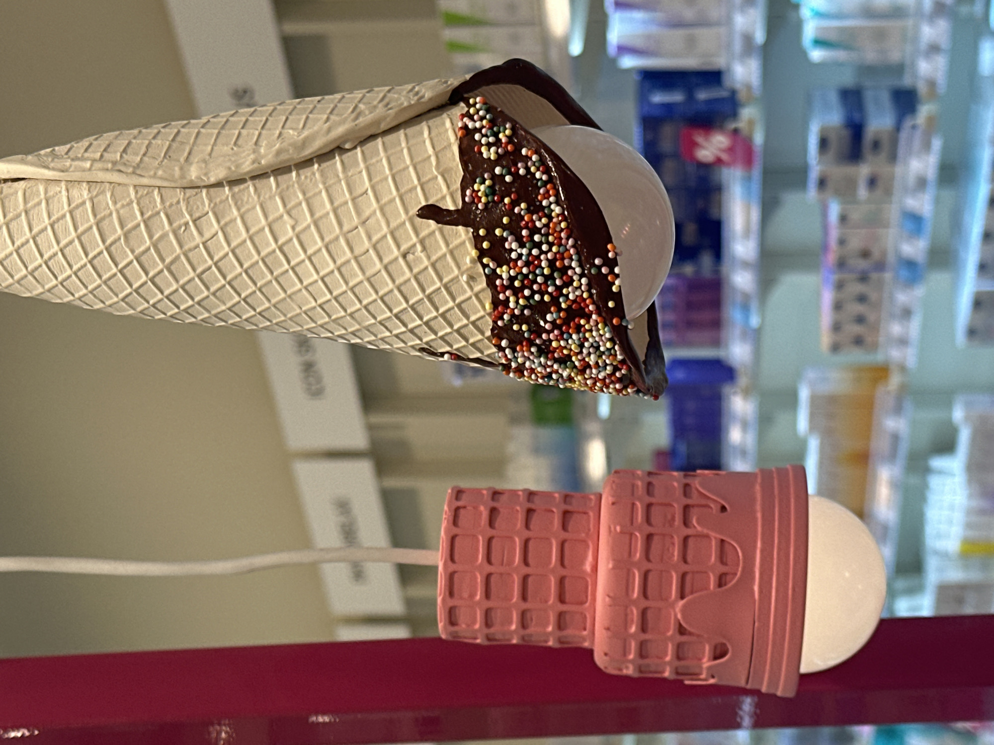The image shows two novelty ice cream cone-shaped lamps. The top lamp is beige with chocolate drizzle and colorful sprinkles, while the bottom lamp is pink with a similar cone pattern. Both lamps have a round, white bulb resembling a scoop of ice cream. A thin white electrical cord extends from each lamp. The background shows shelves stocked with various products, likely in a retail setting. The location is Stavropol, Russia.