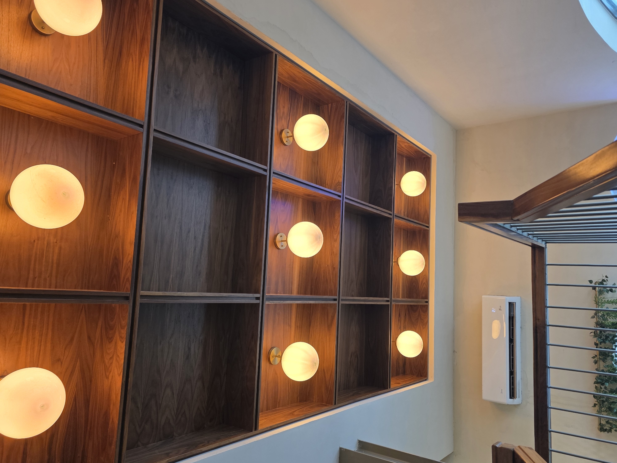 A low-angle interior shot depicts a wall with a large wooden shelving unit featuring illuminated spherical light fixtures. The shelving unit is composed of dark wood, with some sections holding light fixtures and others left empty.
Adjacent to the shelving unit, a wall-mounted air conditioning unit is visible. To the right, a wooden structure partially obscures a view of a window with green foliage visible outside.
The location is likely Tijuana, Mexico, but this information is not directly verifiable from the image content. No people are visible.