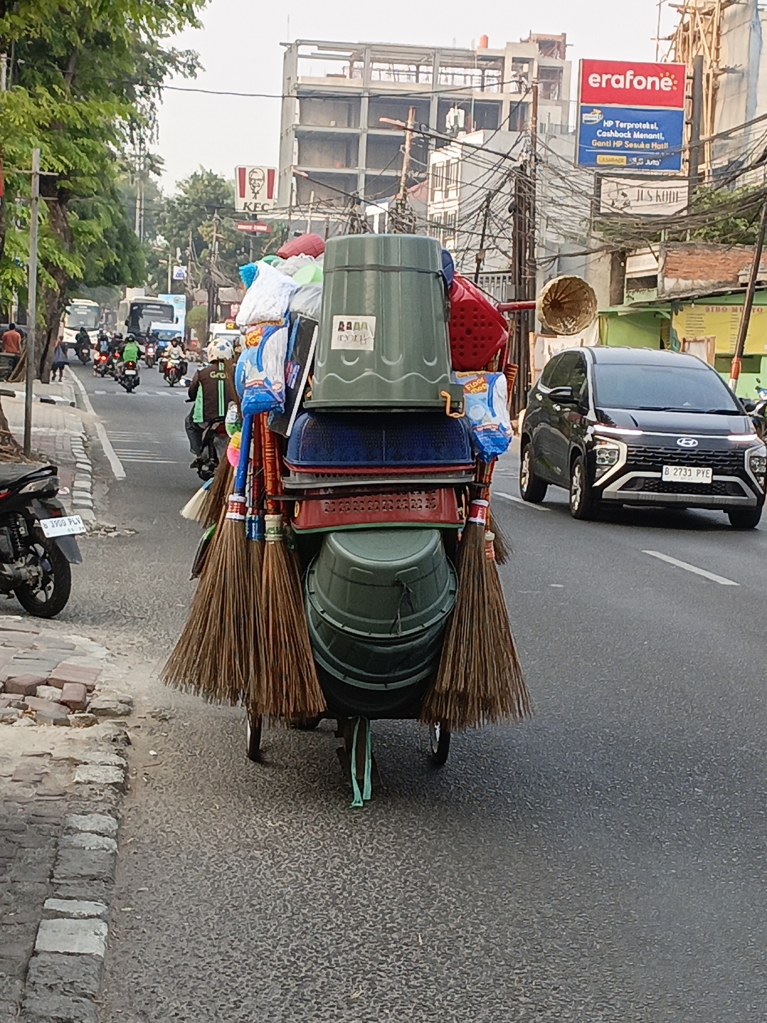 In East Jakarta, Indonesia, a street scene unfolds. A person is pushing a cart loaded with numerous household items, including several large green plastic tubs, brooms, and other various containers and bags. The cart is positioned on the right side of a paved road. To the left of the cart, a sidewalk made of interlocking bricks is visible. Further to the left, a motorcycle is parked on the sidewalk. The street itself is busy with traffic, featuring several motorcycles and a black SUV. In the background, buildings under construction and other structures line the street, along with trees and a KFC sign. A large red and blue advertisement for "erafone" is prominently displayed on one of the buildings. Overhead, a dense network of electrical wires crisscrosses the sky. The lighting suggests it is daytime, possibly late afternoon.