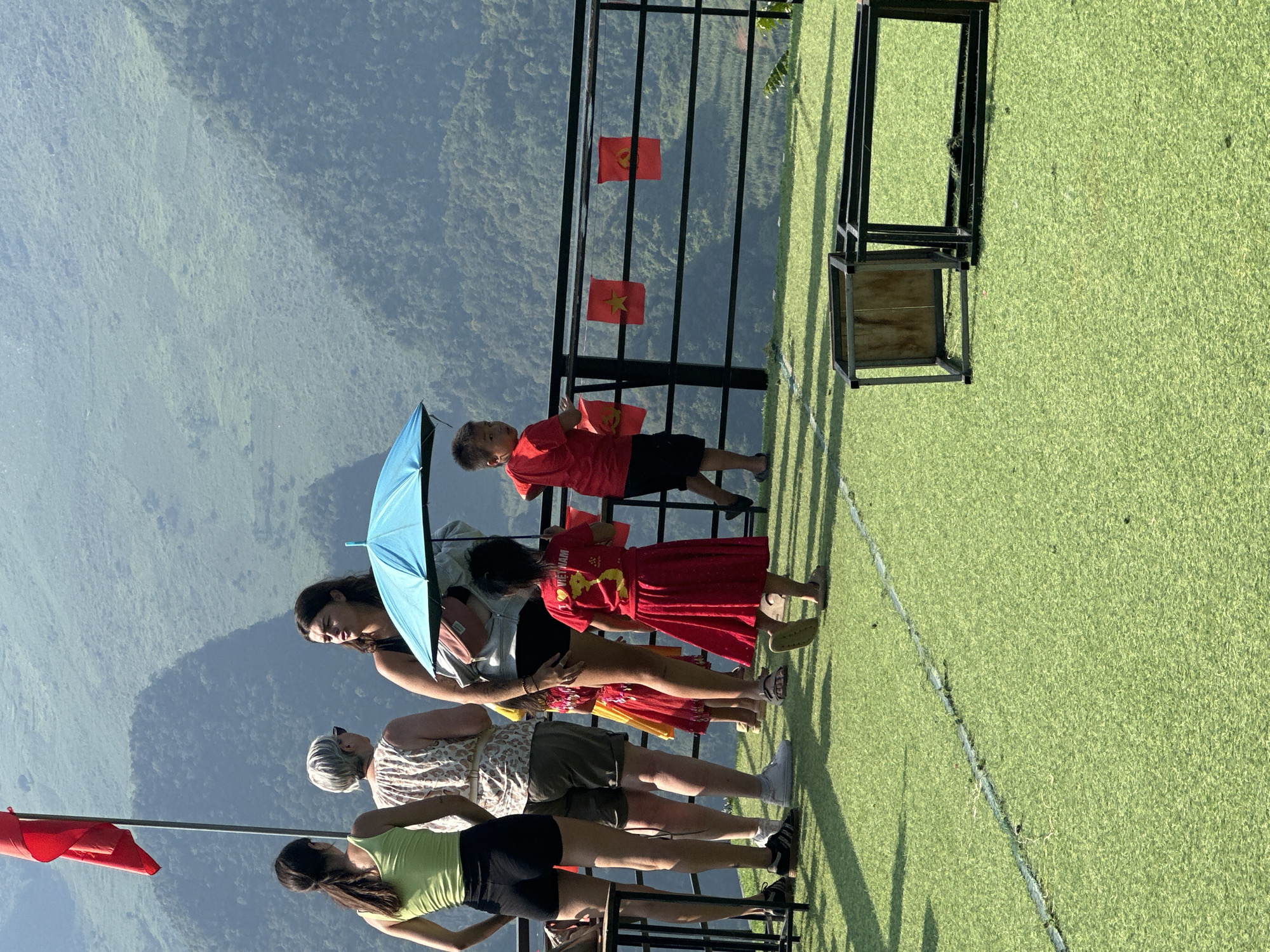 Four women, one child, and a young girl stand along a railing overlooking a valley. The foreground is a green surface with a white line. Behind the railing are small Vietnamese flags attached to the railing. The women are wearing casual clothing. One woman holds a blue umbrella. The child wears a red shirt and the girl wears a red dress. The landscape is a mountainous valley with trees. The location is Đường Thượng Commune, Vietnam.