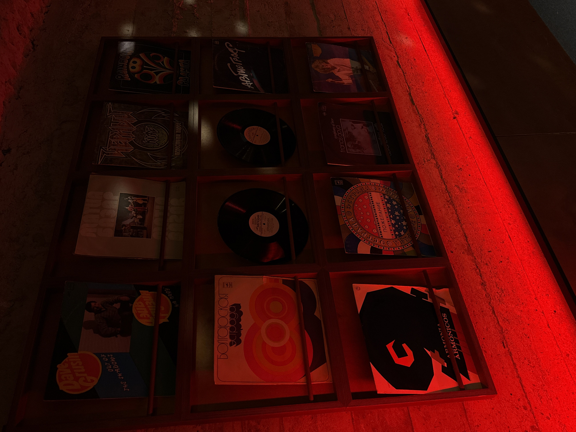 The image shows a display case containing a collection of vinyl records, likely situated in a dimly lit space, possibly a record store or bar. The records are arranged in a grid pattern, with each record occupying its own compartment. The records feature various artists and album covers, including names like Dave Grusin, Rainbow, Francis Goya, and Armonita Armonicos. The albums have Cyrillic text on their covers. The floor and wall next to the case have a red light cast on them. Considering the presence of Cyrillic script on the album covers, and the potential for the display to be in a bar-like setting, it is plausible that this image was taken in Yerevan, Armenia.