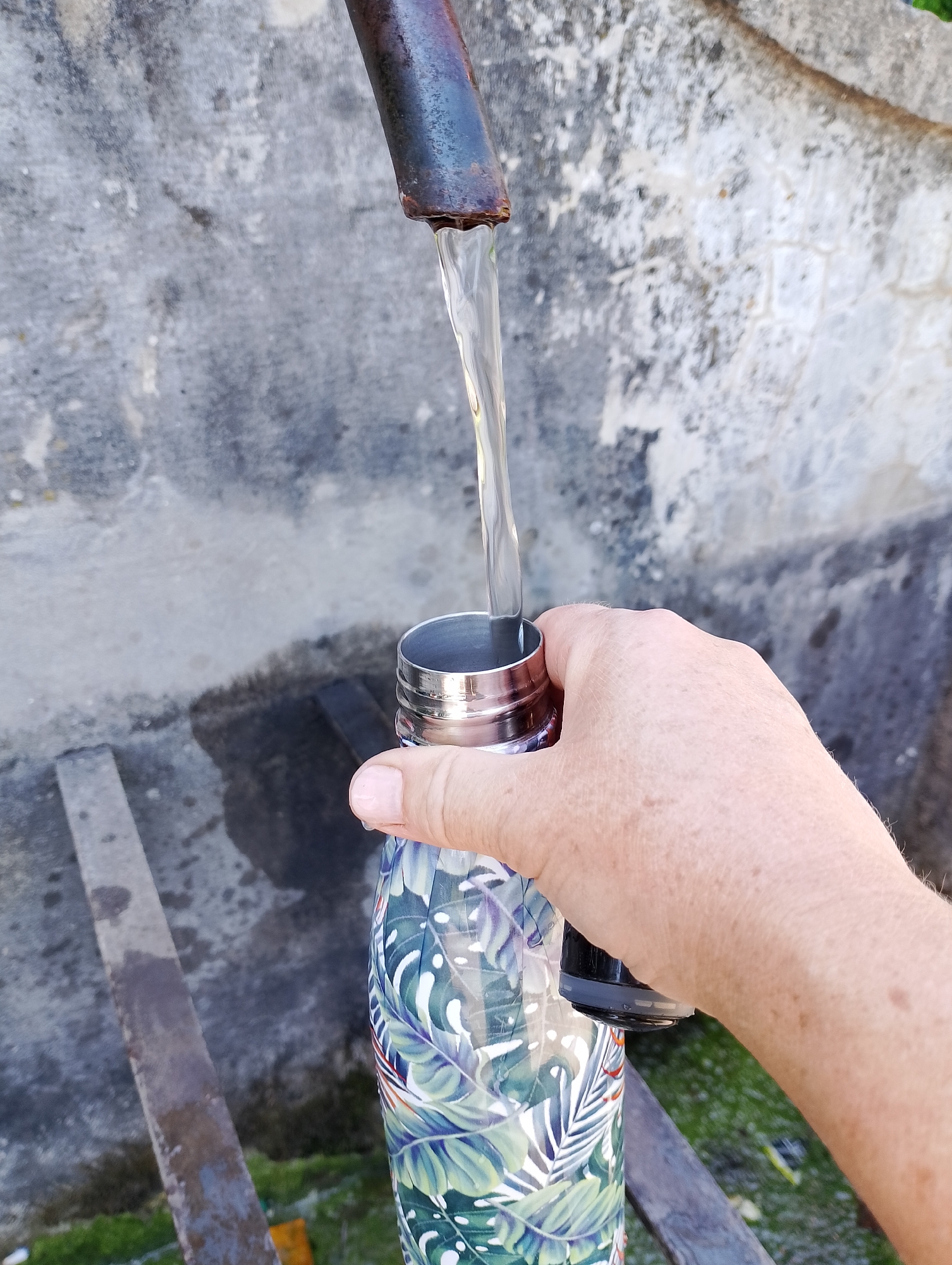 A person's hand holds a reusable water bottle with a tropical leaf pattern, under a rusty metal pipe from which clear water is flowing into the bottle. The background is a textured, weathered concrete wall. The scene is outdoors, possibly in a park or public area.