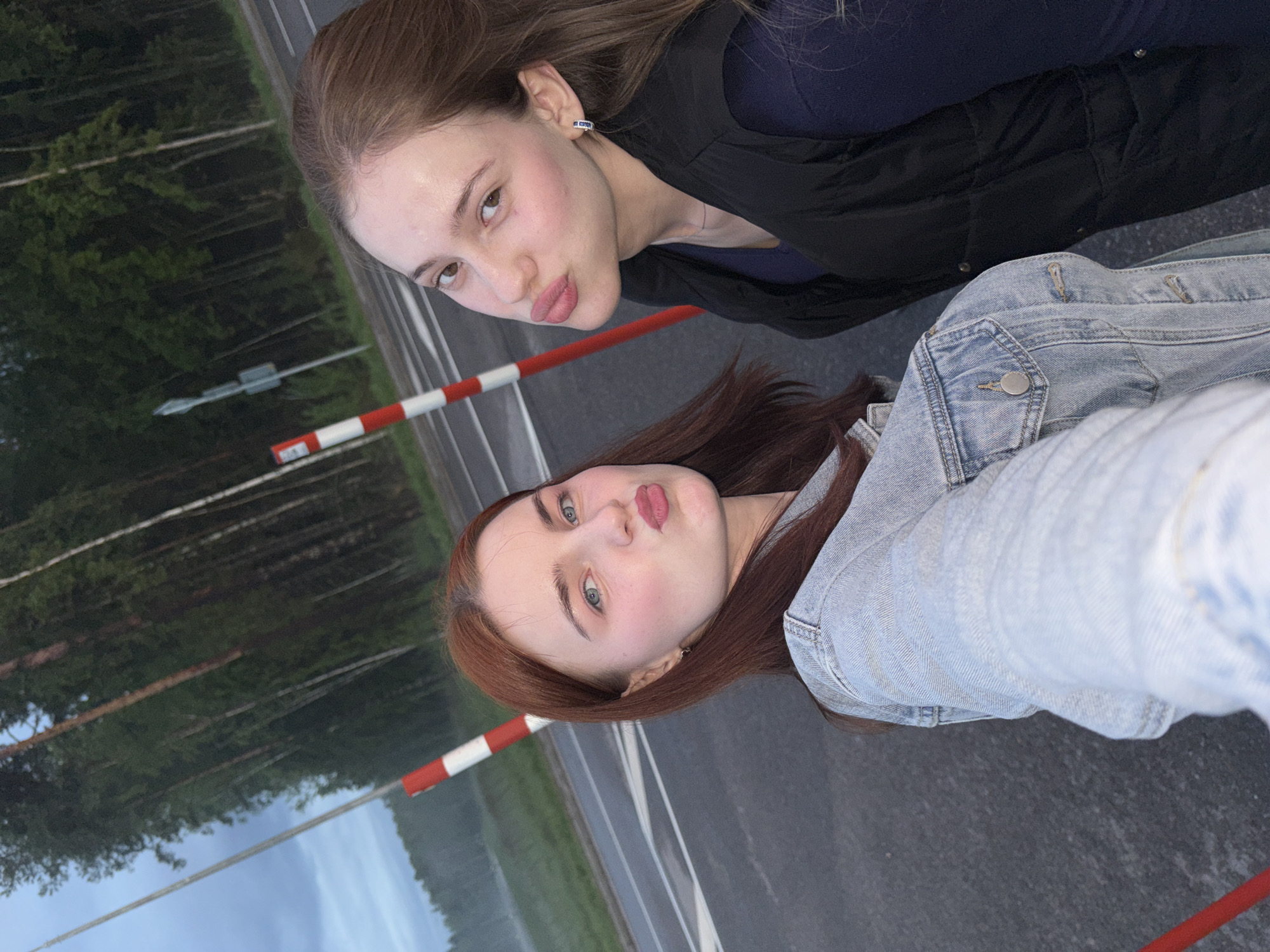 Two young women, posing for a selfie, stand on a paved road with a forest in the background. The woman on the left has auburn hair, blue eyes, and is wearing a light blue denim jacket. She is puckering her lips. The woman on the right has brown hair, brown eyes, and is wearing a dark blue shirt and a black vest. She is also puckering her lips. Red and white striped poles are visible in the background, suggesting they are near a road or a barrier. The sky is overcast. The location is identified as the city of Arfa, Russia.