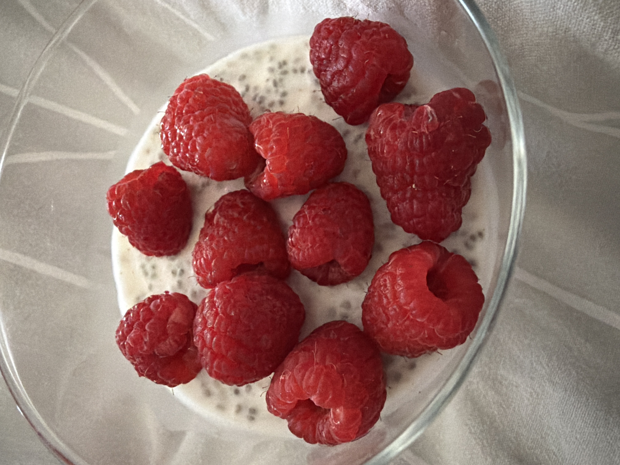 A clear glass bowl contains a creamy white substance speckled with tiny black chia seeds. Multiple bright red raspberries are scattered on top of the mixture. The bowl rests on a light-colored, textured surface, possibly a tablecloth or placemat. The lighting is soft and natural, creating a gentle sheen on the raspberries and the glass. The background is slightly blurred, focusing attention on the food. The scene is an indoor still life, likely a healthy breakfast or dessert, presented in a close-up, top-down perspective. There is no discernible location in Pazardzhik, Bulgaria, or any people present.