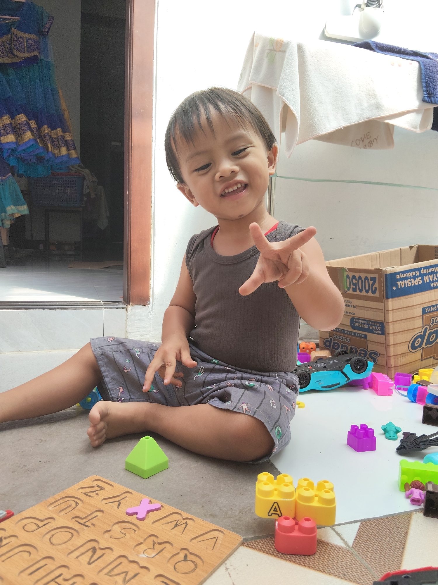 A young boy is sitting on the floor of what appears to be a covered outdoor patio or verandah in a residential setting in the city of Pemalang, Indonesia. The boy is wearing a dark grey sleeveless shirt and grey shorts with a pattern of small figures. He is looking towards the camera with a wide smile and has his right hand raised, with his index and middle fingers extended in a peace sign. Surrounding him are various children's toys, including a wooden alphabet puzzle with some letters missing, colorful building blocks (yellow, red, and purple), a blue toy car, and other plastic toys scattered on the tiled floor. In the background, a doorway leads into a darker interior, with a blue and gold traditional garment hanging on the left. A cardboard box with Indonesian text is visible to the right, along with some folded white cloths hanging on a line. The lighting suggests it is daytime.