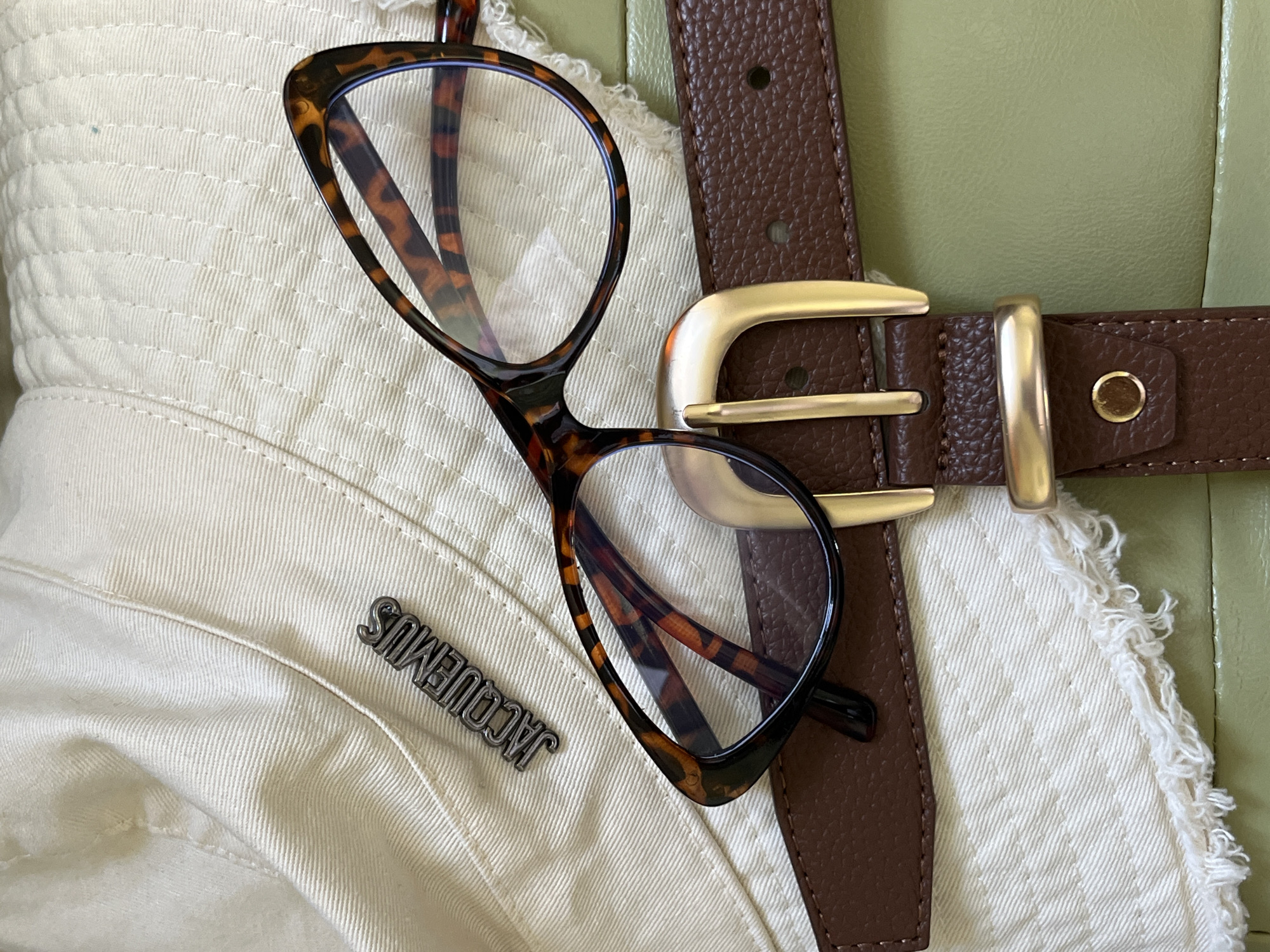 An overhead, close-up shot shows a tortoise-shell patterned pair of glasses, a brown leather belt with a gold buckle, and a cream-colored bucket hat with a silver "JACQUEMUS" logo. The items are arranged on a light green surface, possibly a sofa or a piece of furniture. The hat has frayed edges, suggesting it might be a denim or canvas material. The belt is textured and has a classic rectangular buckle. The glasses are cat-eye shaped and have a bold, thick frame. The lighting is bright and appears to be natural daylight. The scene is static and there are no people visible. The image does not provide any information to suggest it is located in Starobaltachevo, Russia.