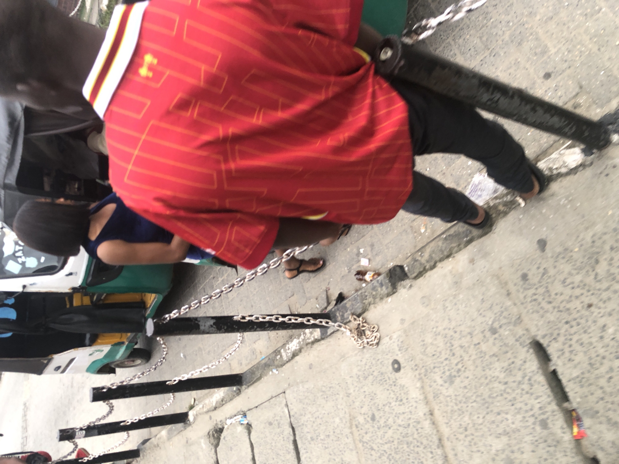 An over-the-shoulder medium shot captures a young Black man from the waist down, walking away from the camera. He wears a bright red polo shirt with yellow stripes and a yellow crest on the right shoulder. His dark pants and brown sandals are visible. A chain fence runs diagonally across the foreground, with black posts and silver links. To the left of the fence, a grey concrete sidewalk is visible, and beyond that, a dirt shoulder with scattered debris. In the background, a white, green, and yellow motorized tricycle, commonly known as an "Okada" or "Keke Napep," is parked on the street. A Black woman in a dark blue sleeveless dress is partially visible next to the tricycle, facing away from the camera. The scene appears to be a street in Ado, Nigeria, likely a commercial area with pedestrian traffic.