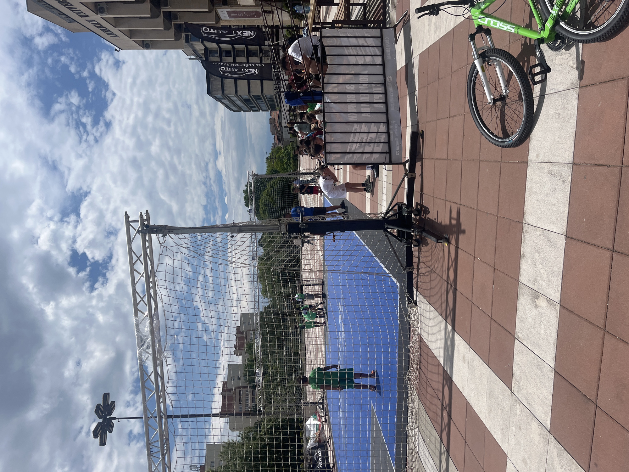 A net-enclosed sports court occupies an outdoor public area in Blagoevgrad, Bulgaria. The court has a blue playing surface. Several individuals in green sports uniforms are on the court. Spectators are seated in a raised structure next to the court. A green bicycle is parked nearby on a red and white tiled surface. Buildings are in the background and the sky is partly cloudy. The location is likely a street, as indicated by the street lamp. Banners with the text "NEXT AUTO" in Bulgarian are displayed on buildings.