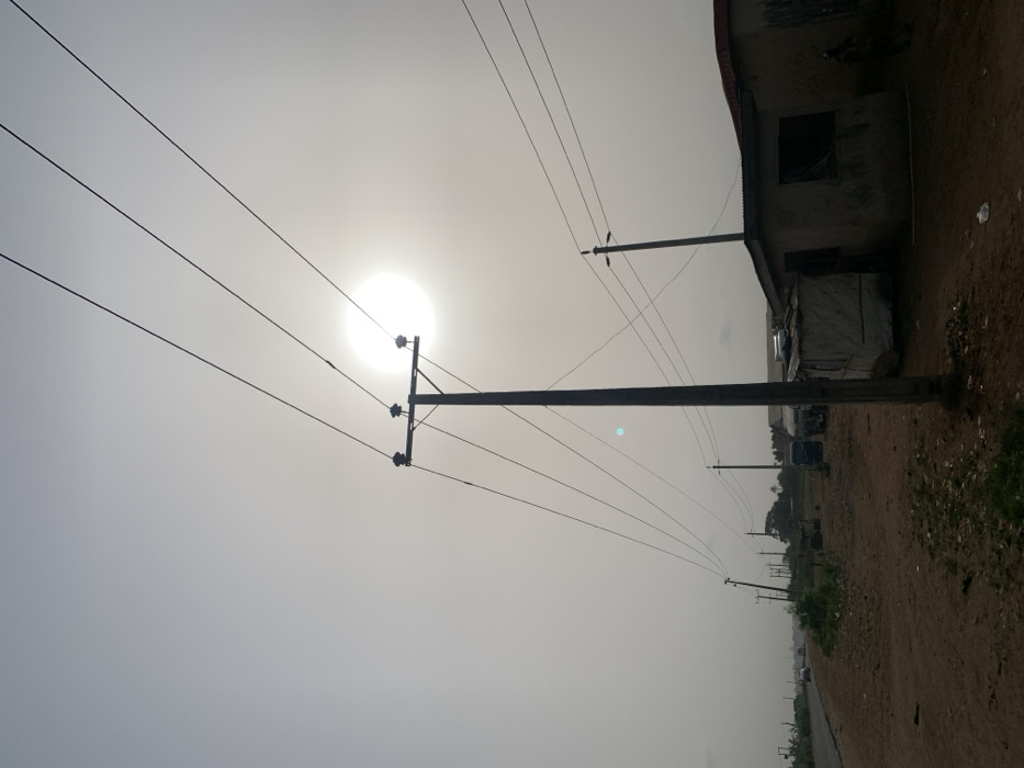 The image captures an outdoor scene dominated by utility poles and power lines against a bright, hazy sky. The overall impression is one of strong backlighting.
In the upper-left quadrant of the frame, a brilliant, almost perfectly circular and intensely white light source, presumed to be the sun, shines through a thick haze. The sky around it is a very light gray or off-white, indicating either heavy cloud cover or atmospheric haze, which diffuses the sunlight.
Several utility poles are visible. A large, dark-colored pole stands prominently in the mid-left portion of the image, extending from near the bottom edge upwards. It is seen in silhouette against the bright sky, clearly showing insulators and cross-arms from which multiple power lines extend. These thin, dark power lines crisscross the upper and middle sections of the sky, running horizontally across the frame and connecting to other poles. Another utility pole is situated further back and to the right, and more poles can be seen receding into the distance along a path.
On the far right side of the image, a structure, likely a building, is partially visible. It appears dark, possibly due to being unpainted or in shadow, with a rough texture. A corrugated metal roof is distinguishable at the top edge of the building. There's a dark, rectangular opening that might be a window or doorway. Below this main structure, a lighter-colored, possibly temporary shelter or stall, perhaps covered with a tarp, is also present.
The ground along the right side of the image is a dirt road or path, uneven and appearing somewhat barren. Sparse patches of green vegetation or grass can be seen along the edge of the path, particularly further into the background and on the extreme right. Some debris is scattered on the ground near the building. In the distance, vague shapes of what might be vehicles or other structures are discernible but indistinct due to the distance and atmospheric conditions. A faint blue-green lens flare is visible in the middle-right area of the sky, consistent with the camera pointing directly at the sun.
The intense brightness of the sun and the hazy conditions suggest it could be midday or early afternoon. There is no visible text in the image.