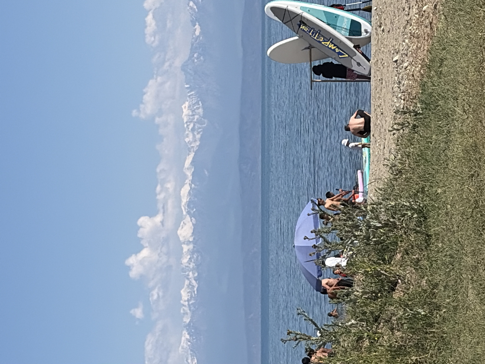 The image depicts a sunny day at a beach with a group of people enjoying the water. In the foreground, lush green vegetation lines the sandy shore. Several individuals are visible, some lounging on the beach, others in the water. One person is lying on the sand, facing away from the camera, wearing black shorts. Another person, wearing a white hat and black shorts, sits by the water.
In the water, a large blue umbrella provides shade. Several people are gathered around the umbrella and on paddleboards. Two paddleboards are propped up on the shore, one white with teal accents and the other mostly white. The word "COMPETITION" is visible in yellow lettering on one of the paddleboards. The water is a deep blue, and in the distance, snow-capped mountains rise majestically against a clear blue sky with scattered white clouds. The overall impression is a leisurely summer day by the water.