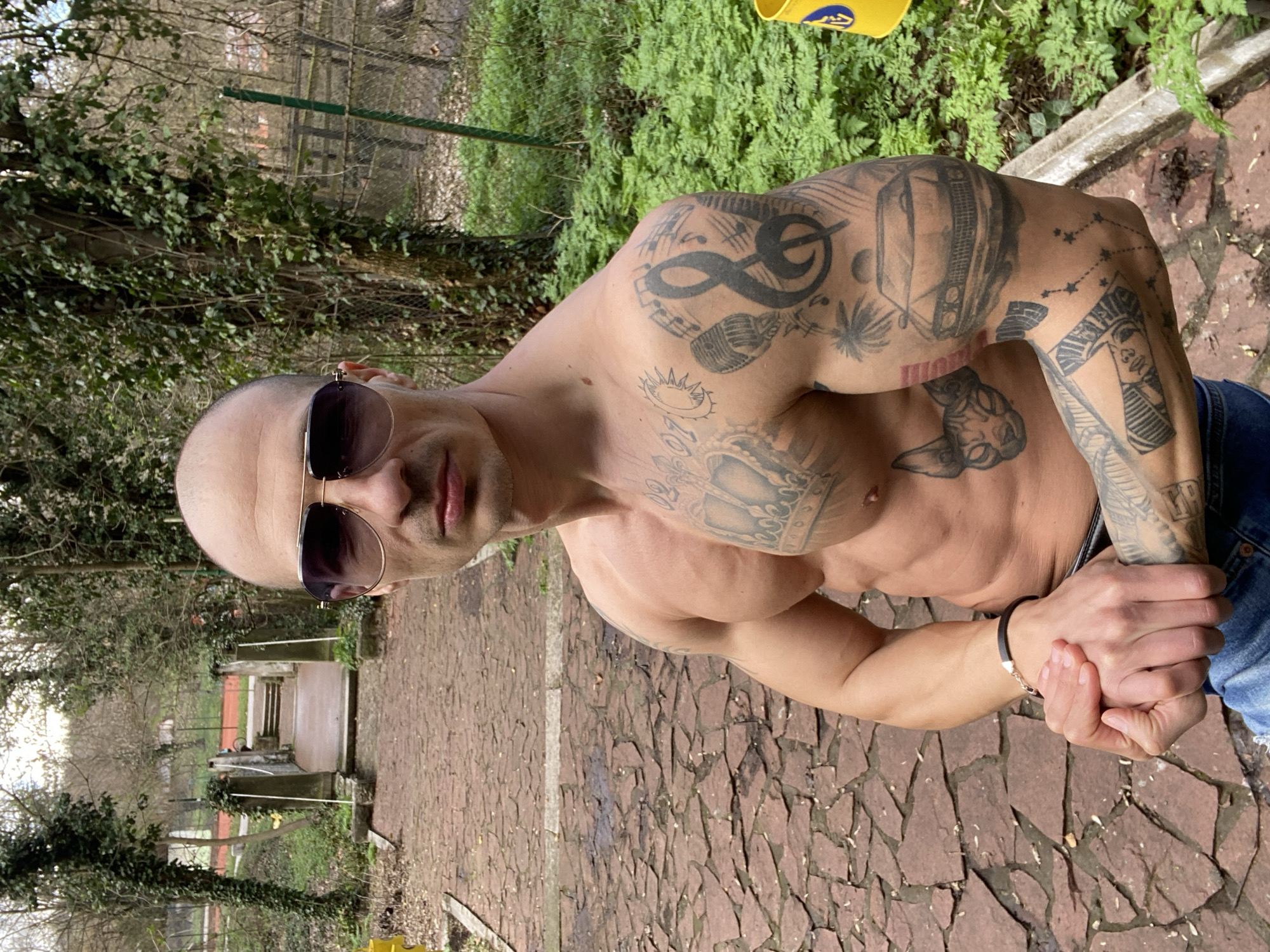 A bald, muscular male subject stands shirtless on a damp, reddish-brown irregularly paved pathway. He wears dark sunglasses with gold frames and partially visible blue jeans. His hands are clasped together at waist height, and a dark bracelet with a silver clasp is on his left wrist. The subject is heavily tattooed, featuring a large crown with "07-07" and a sun on his chest, a Sphynx cat outline on his abdomen, and a treble clef with musical notes, a microphone, palm trees, a classic car, and a star constellation on his right arm.
The background consists of dense green foliage and ivy-covered trees. In the mid-ground, a stone bridge with railings crosses a lower section of the park. Beyond the bridge, a red clay tennis court with white lines and a net is visible, partially obscured by a green wire fence. A yellow plastic container is partially visible on the far right. This location is consistent with a recreational area within Kaylaka Park in Pleven, Bulgaria.