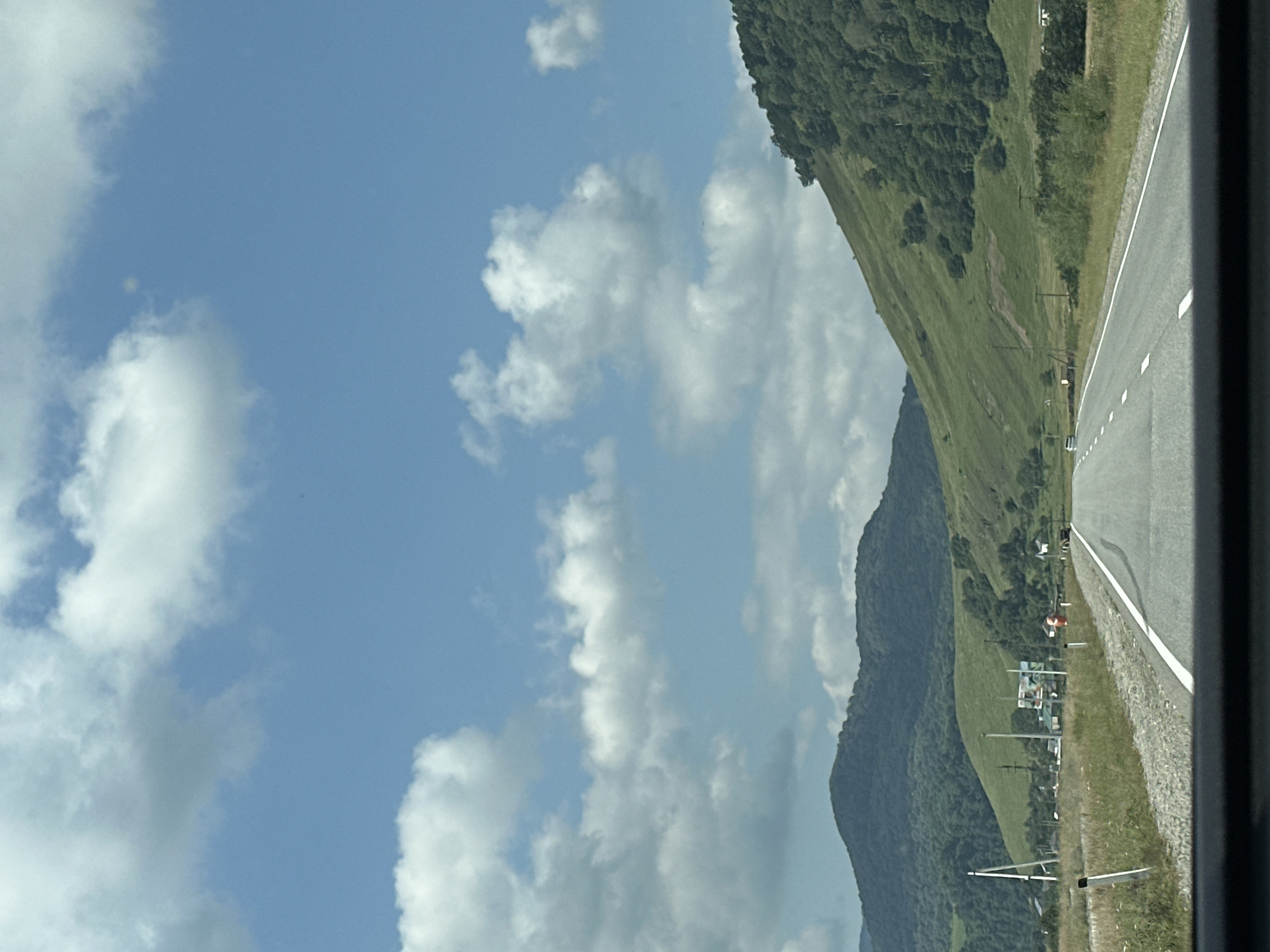The image depicts a road leading through a rural landscape with rolling green hills under a cloudy blue sky. On the right side of the road, a steep, tree-covered hillside dominates the scene, with patches of grass and some smaller trees visible on its lower slopes. The road itself is paved with white lane markings. To the left of the road, there is a grassy verge, followed by fields with scattered vegetation. In the distance, there are more hills covered in trees and a few small buildings, suggesting a rural settlement. There are utility poles and traffic signs along the roadside. The lower part of the image shows the dashboard of a vehicle, indicating the photo was taken from inside a car. The scene is devoid of people. This appears to be a view of the road on the outskirts of Nizhnyaya Yermolovka, Russia, characterized by its natural, mountainous terrain.