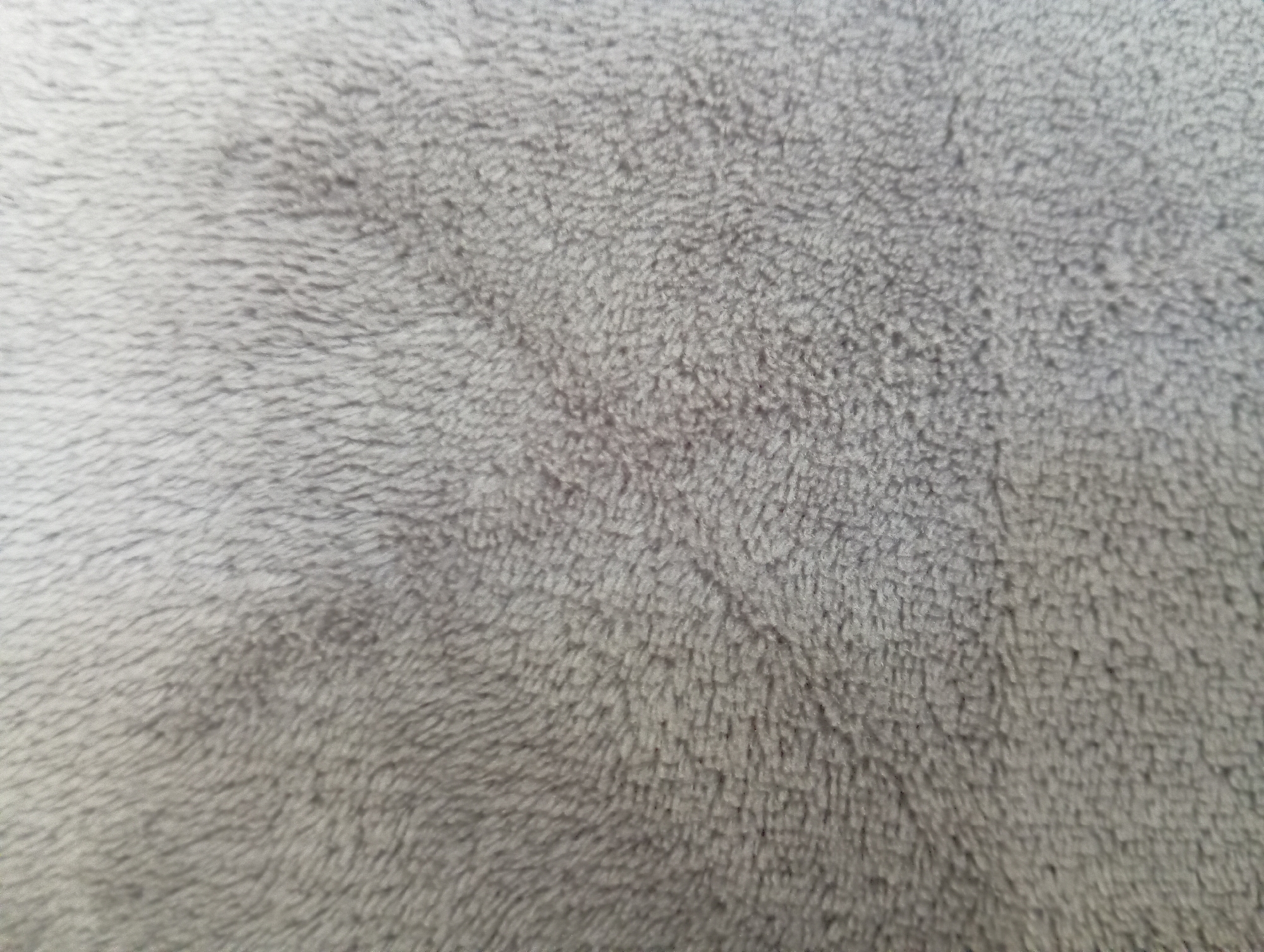 The image is a close-up, overhead shot of a gray, plush fabric. The texture is soft and appears to be a type of faux fur or fleece. The fibers are short and densely packed, creating a slightly uneven surface with subtle variations in light and shadow due to the direction of the pile. There are no people, objects, actions, or interactions visible. The image depicts only the fabric. The provided information about Hlynytsi, Ukraine is not reflected in the visual content of the image.