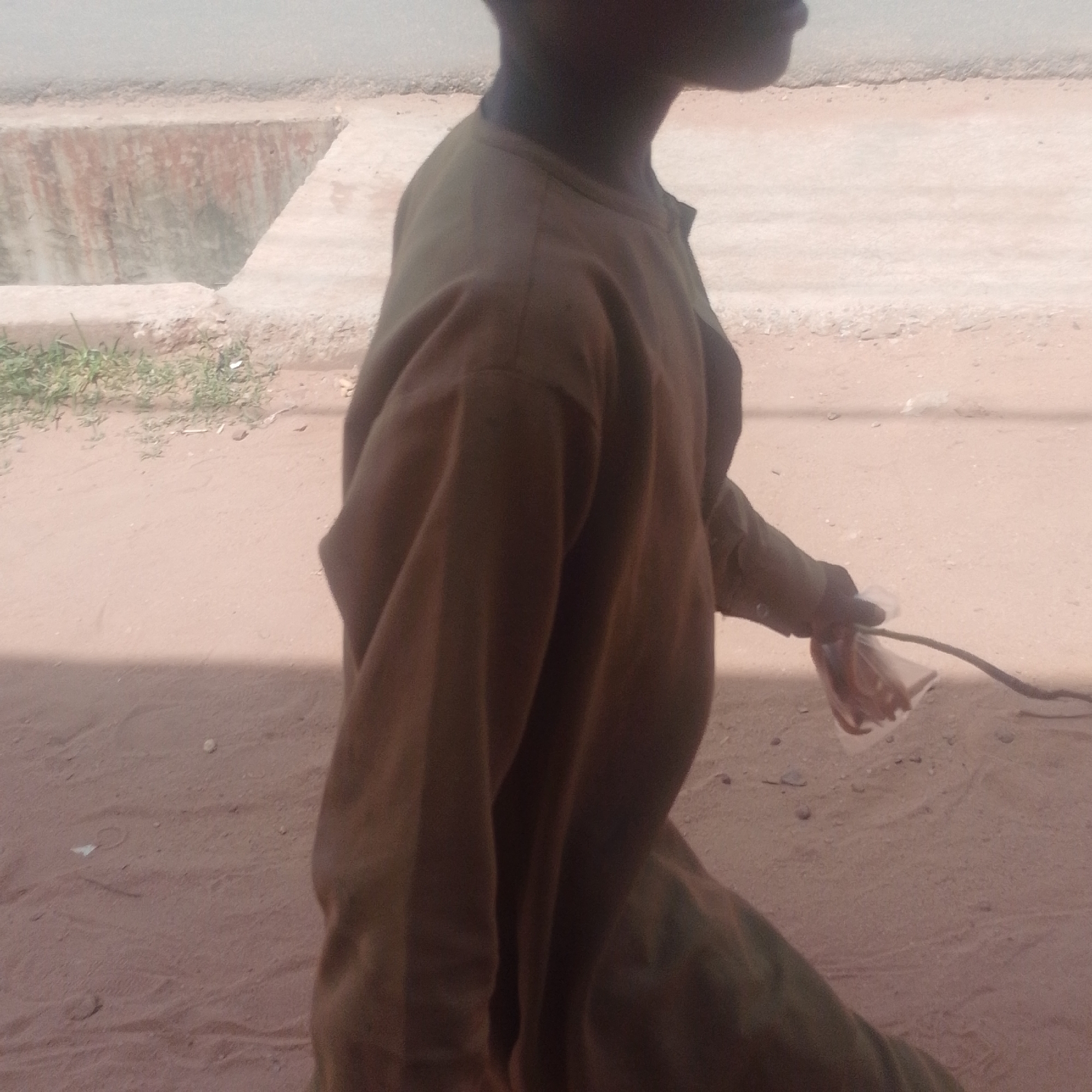 The image depicts a person walking on a sandy, unpaved street in Gombe, Nigeria. The individual is wearing a loose-fitting, long-sleeved brown garment. Their head and upper body are visible, in profile, facing towards the right of the frame. They are holding a clear plastic bag in their right hand, which appears to contain some red-colored items, and a dark, thin cable or rope is emerging from the bag and extending towards the right edge of the image. The ground is composed of light brown sand, with some small stones and sparse patches of green vegetation visible. In the background, there is a concrete structure that resembles a drainage ditch or culvert, with reddish staining on its interior walls. The overall environment suggests a rural or semi-urban setting within Gombe.