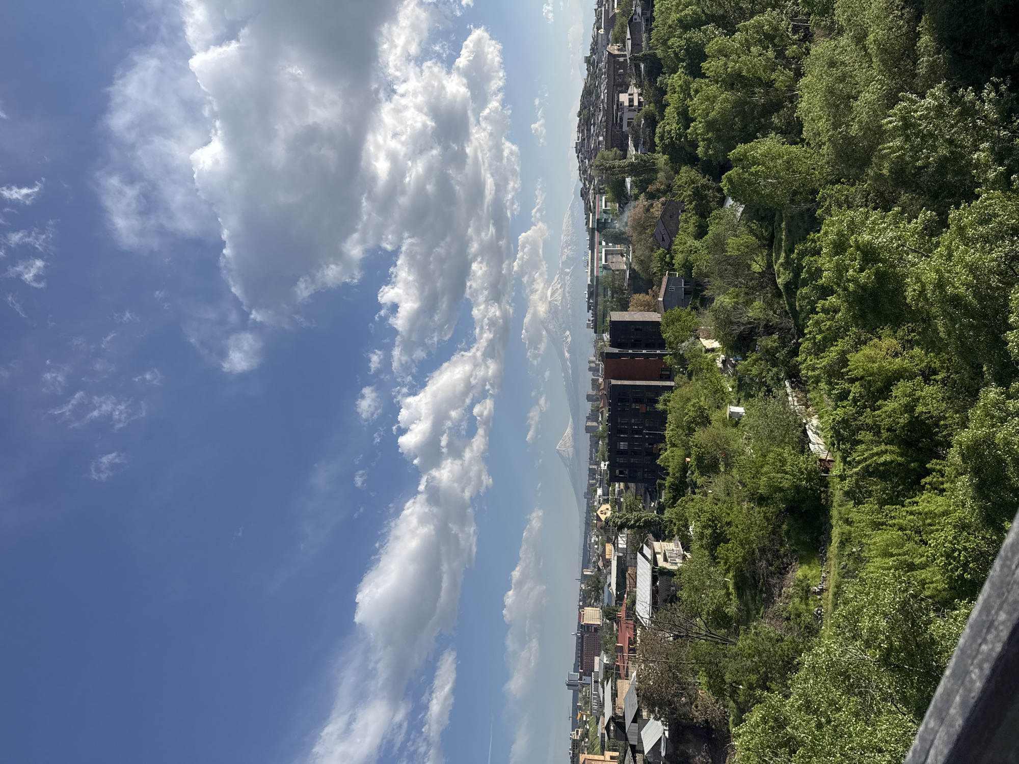 The image captures a panoramic view of Yerevan, Armenia, under a partly cloudy sky. The cityscape unfolds with buildings of varying heights and architectural styles nestled among lush greenery. The sky is dominated by large, billowing white clouds contrasting with patches of clear blue. In the distance, the faint outline of Mount Ararat can be seen through the haze. The foreground includes a partial view of a wooden structure. The perspective suggests the photo was taken from an elevated vantage point, likely overlooking a residential or urban area.