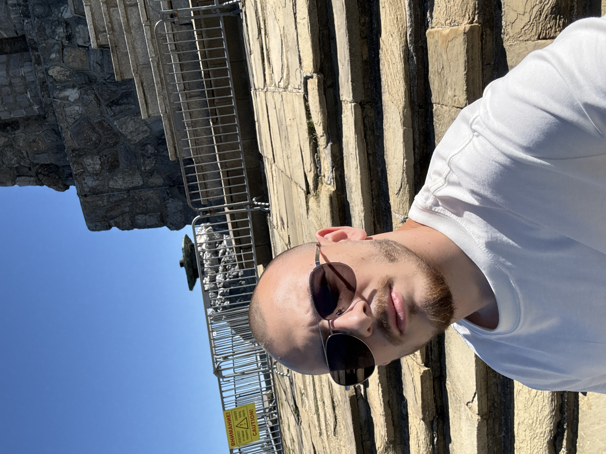In the city of Shipka, Bulgaria, a young man with a shaved head and a goatee is captured in a selfie. He is wearing a white t-shirt and sunglasses, with a slight smile on his face. The man is seated on rough, weathered stone steps that appear to be part of a monument or historical site. In the background, a metal fence separates the viewer from a stone structure and a clear blue sky. A yellow caution sign with text in both Cyrillic and English is attached to the fence. The overall environment suggests an outdoor location on a sunny day, possibly a tourist attraction.
