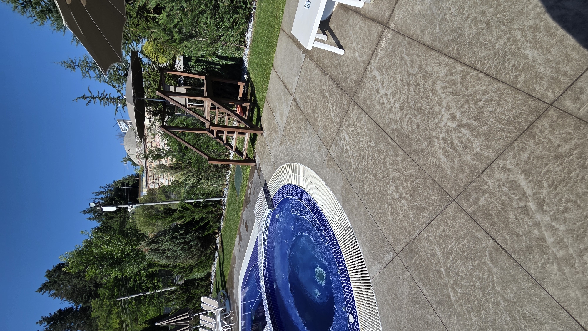 The image depicts an outdoor scene in Bania, Bulgaria, likely a resort or hotel area, featuring a swimming pool and hot tub. The pool is circular and filled with bright blue water, tiled with smaller blue and white mosaic tiles around its rim. White grating surrounds the pool's edge. Adjacent to the pool is a paved patio area with large, textured, beige tiles.
To the right of the pool, a wooden structure with stairs leads up to a raised platform, possibly a lifeguard stand or viewing deck. Two large, neutral-colored umbrellas are positioned nearby, providing shade. A white plastic lounge chair and a small white side table are visible on the patio.
The background is lush and green, with numerous evergreen trees and bushes. A stone wall with some decorative elements runs horizontally behind the trees. In the distance, a dome-shaped structure, possibly a part of a historical building or a spa, is visible against a clear blue sky. Several streetlights and security cameras are mounted on poles amidst the foliage. The overall impression is a well-maintained outdoor recreational space during daylight hours. There are no people visible in the image.