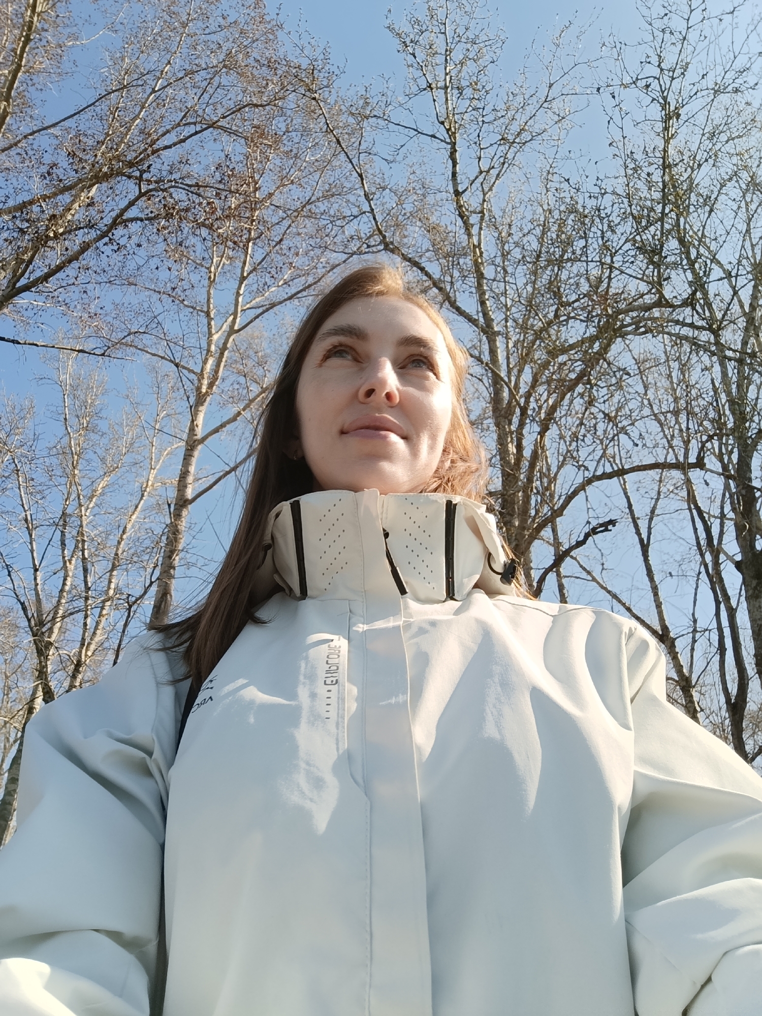 A low-angle, full-shot photograph shows a young woman in a white jacket looking upward. The woman has fair skin, brown hair, and is facing slightly to the right. The jacket has a distinctive high collar with dark trim. Behind the woman is a sky with bare-branched trees, typical of a late fall or winter scene. The sky is pale blue. The photo appears to have been taken in the city of Chaikovsky, Russia, based on the location metadata. The focus is primarily on the woman and the immediate background.