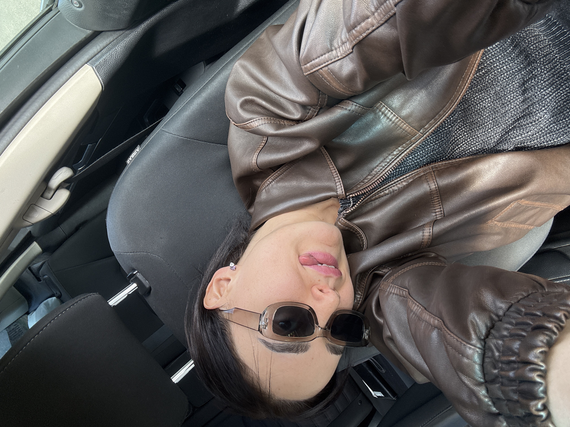 A person wearing a brown leather jacket and sunglasses sits in the driver's seat of a vehicle. The person's head is tilted backward, and their tongue is slightly visible. The interior of the vehicle is visible, including the seat and dashboard. The location of the image is unknown, but it is not in Tbilisi, Georgia, as there is no information within the picture to connect the location.