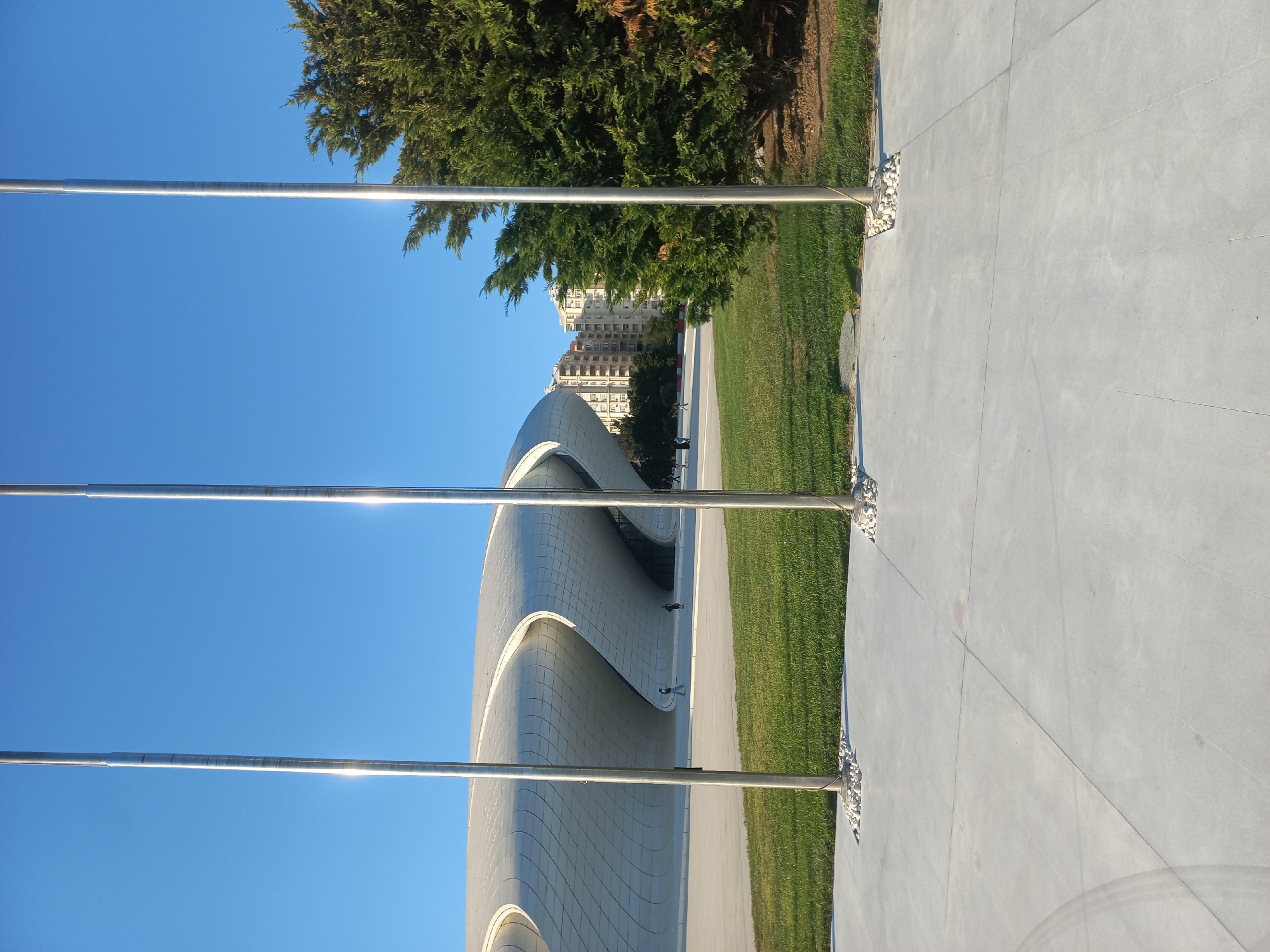 The image depicts the Heydar Aliyev Center in Baku, Azerbaijan, a striking example of modern architecture. Three tall, metallic flagpoles stand in the foreground, casting reflections on their polished surfaces due to the bright sunlight. Behind the flagpoles, a vast expanse of bright green, well-maintained grass stretches out.
Further back, the iconic Heydar Aliyev Center's white, flowing structure is visible, characterized by its undulating curves and seamless design. The building appears to be a cultural center or museum, with its unique architectural style standing out against the clear blue sky. In the background, several multi-story residential buildings, typical of urban environments, are present. A large, dense evergreen tree is situated on the right side of the frame, adding a natural element to the scene.
A few individuals are seen walking in the middle ground, near the Heydar Aliyev Center, indicating that the area is open to the public and likely a popular spot for visitors and locals. The ground in the foreground is paved with light gray concrete slabs, marked by clean grout lines. The overall impression is one of a modern, well-organized public space bathed in sunlight, showcasing contemporary architecture and urban landscaping.