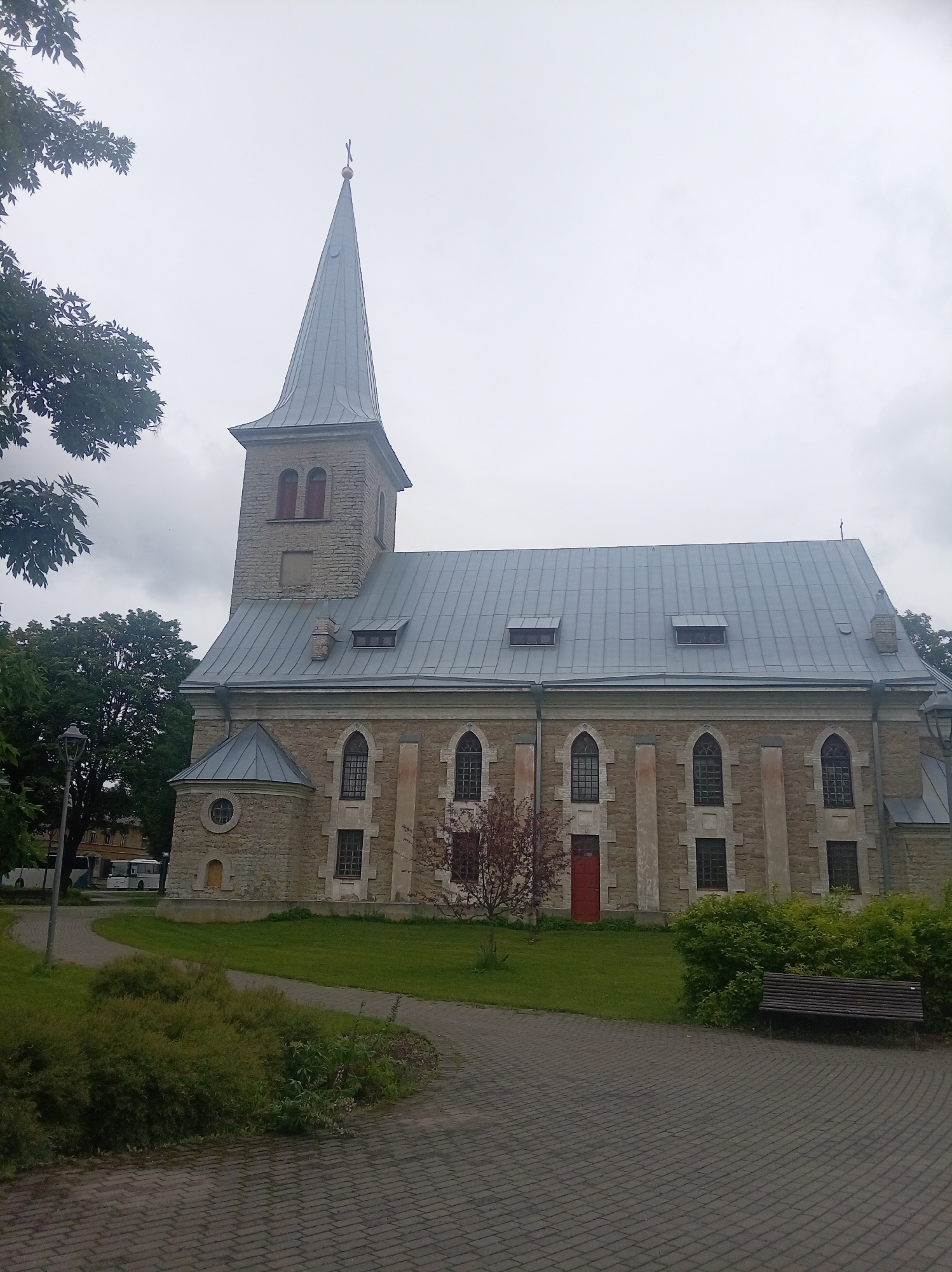 The image shows the Tapa Evangelical Lutheran Church in Tapa, Estonia. The church is a stone building with a tall, pointed spire topped with a cross. The roof is made of gray metal. The church has several arched windows with dark frames and red doors. In front of the church, there is a paved walkway and a patch of grass with a bench. Trees and bushes are also visible in the scene. The sky is overcast.