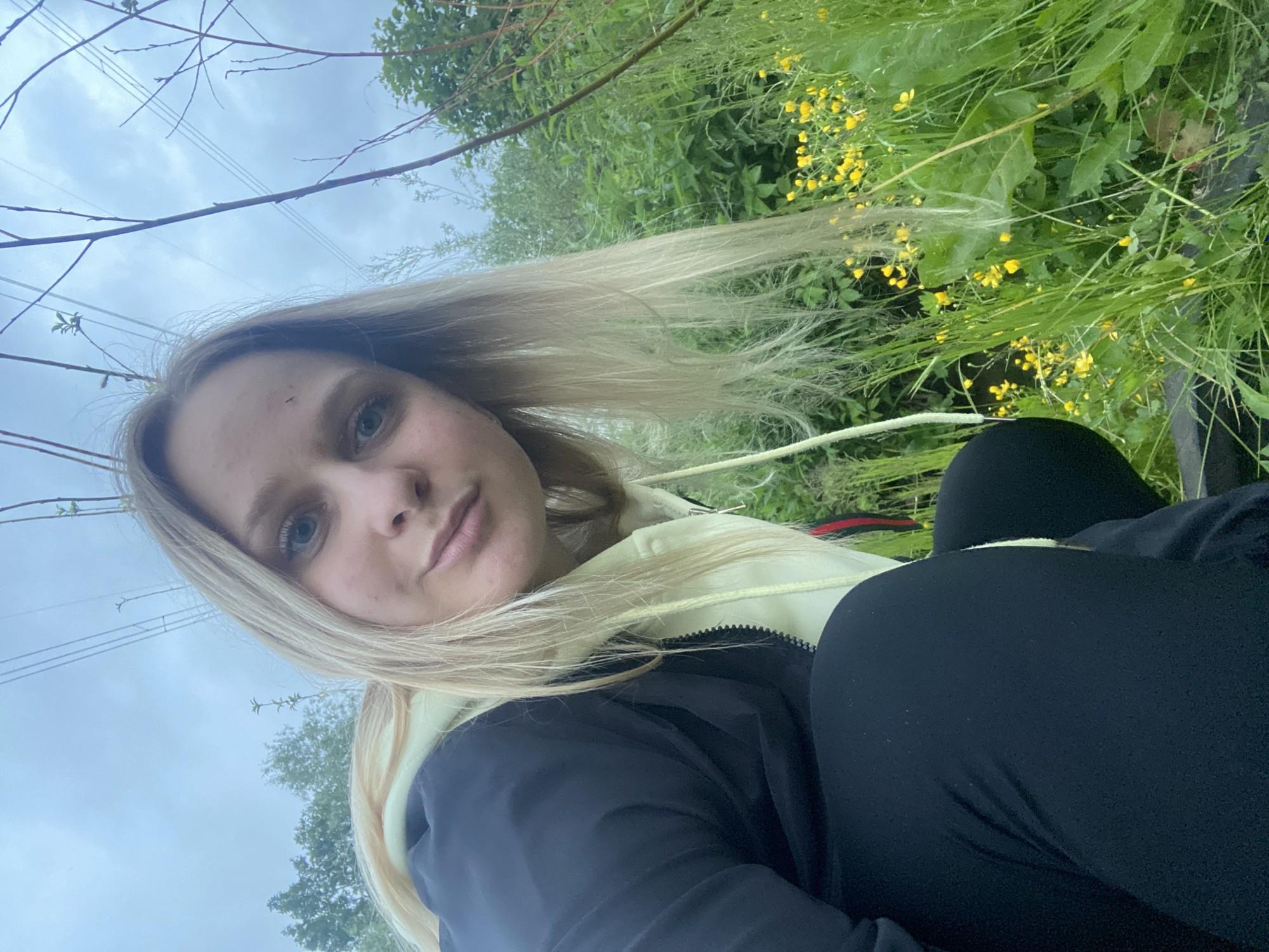 A young adult female with long blonde hair and light-colored eyes is captured in a close-up, low-angle outdoor portrait. She is positioned in a crouched or squatting posture, looking directly towards the camera with a neutral expression. She is wearing a black zip-up jacket over a light yellow or lime green hooded sweatshirt with visible drawstrings. Her lower body is clad in dark-colored, form-fitting pants or leggings. The background features a dense growth of green foliage, including tall grass, bushes, and distant trees, indicative of an overgrown or natural area. Small clusters of bright yellow flowers are visible amidst the green vegetation on the right side of the frame. The sky above is overcast, displaying light gray and blue tones. High-voltage power lines are distinctly visible spanning across the upper portion of the sky, intersecting with slender, bare tree branches. The location is an outdoor, natural setting, unidentifiable to a specific city or region.