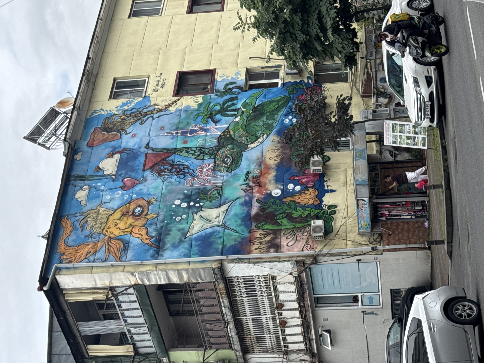 The image shows a colorful mural painted on the side of a multi-story building in Batumi, Georgia. The mural depicts an underwater scene with various marine life, including a large goldfish, a sea turtle, and jellyfish, as well as some man-made structures resembling skyscrapers and a Ferris wheel. The building itself appears to be aged, with balconies, windows, and air conditioning units visible. In the foreground, a street scene unfolds with cars, including a white sedan and a silver hatchback, and a person riding a scooter. The sky is overcast. Signs are visible on the building, including one that appears to be for a dental clinic and another advertising tours in Georgia.