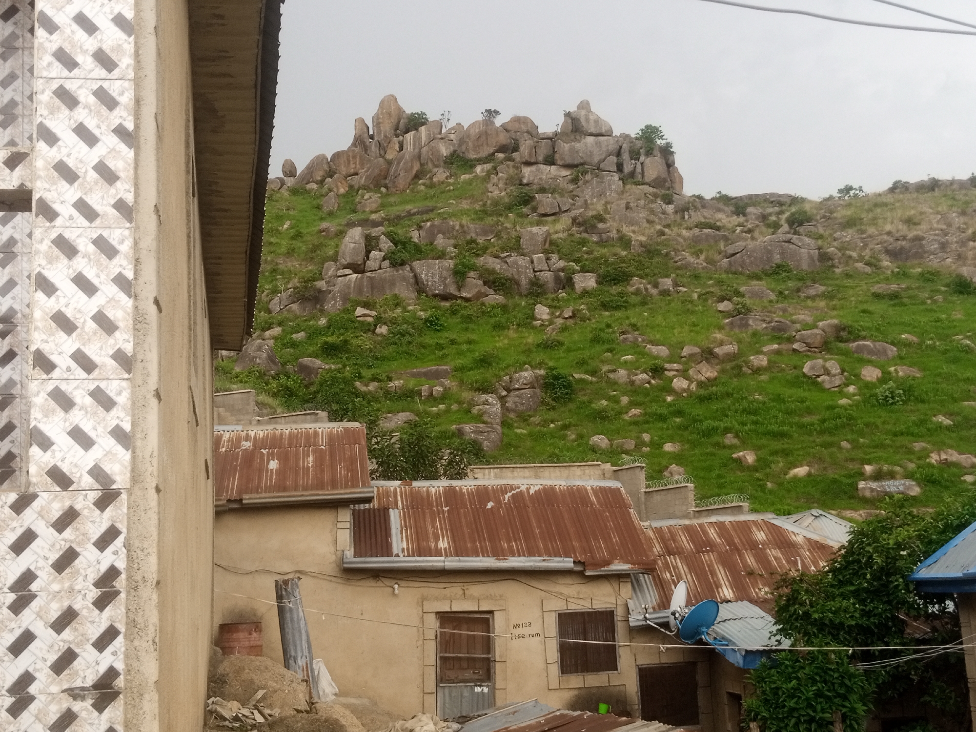 The image shows a cluster of buildings with rusty corrugated metal roofs, situated at the base of a rocky, grass-covered hill. The foreground is dominated by a tiled wall on the left and the side of a building. The buildings appear to be low-rise, with at least one featuring barred windows and a satellite dish. The hill behind the buildings is strewn with large rocks and sparse vegetation, leading up to a more densely packed rocky outcrop at the crest. The sky is overcast. The scene suggests a rural or semi-urban environment in Rukuba Road, Nigeria, characterized by simple architecture and a rugged natural landscape.